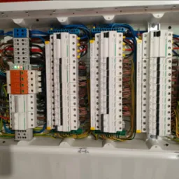 Rozdzielnica elektryczna w białej obudowie z zainstalowanymi bezpiecznikami, przewodami i listwami zaciskowymi. Widoczne oznaczenia i etykiety.