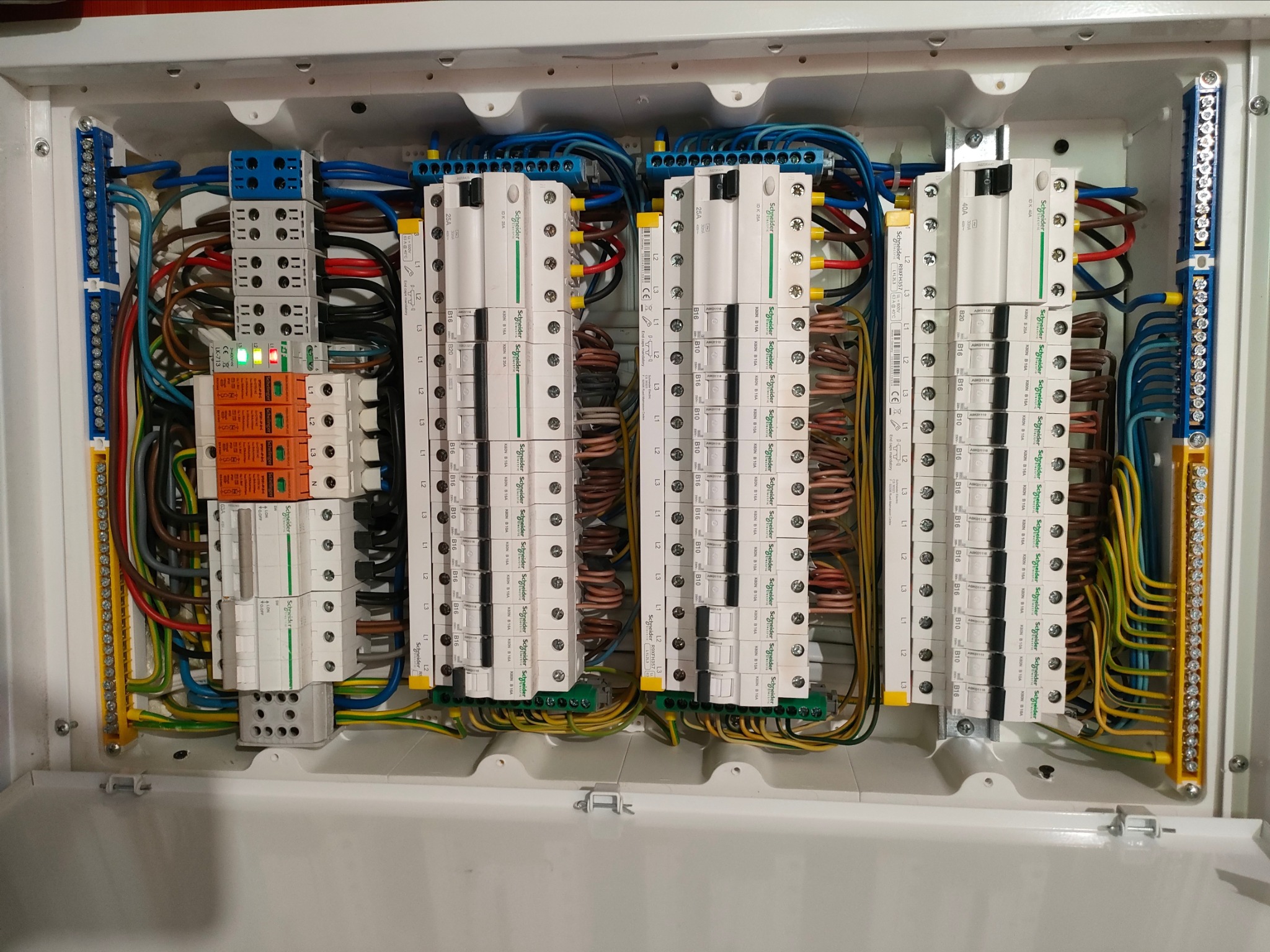 Rozdzielnica elektryczna w białej obudowie z zainstalowanymi bezpiecznikami, przewodami i listwami zaciskowymi. Widoczne oznaczenia i etykiety.