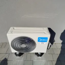 Biała, zewnętrzna jednostka klimatyzacji Midea zamontowana na szarym bruku, przy szarej ścianie. Widoczne śruby mocujące i cień rzucany na ścianę.