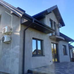 Kowa-San - Szary dom jednorodzinny z zamontowanymi klimatyzatorami na elewacji i oknami z ciemną ramą. Widoczne rynny i fragment tarasu z kostki brukowej. Błękitne niebo w tle.