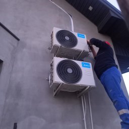 Kowa-San - Dwa klimatyzatory Midea zamontowane na szarej elewacji budynku w Radzyminie. Instalator w niebieskim kombinezonie i czerwonym kasku podczas montażu.