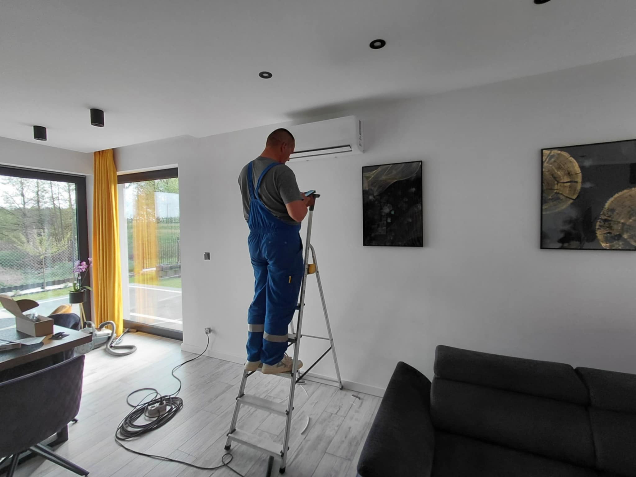 Instalator na drabinie montuje klimatyzator ścienny w nowoczesnym salonie. Widoczne narzędzia, okno z żółtą zasłoną i obrazy na ścianie.