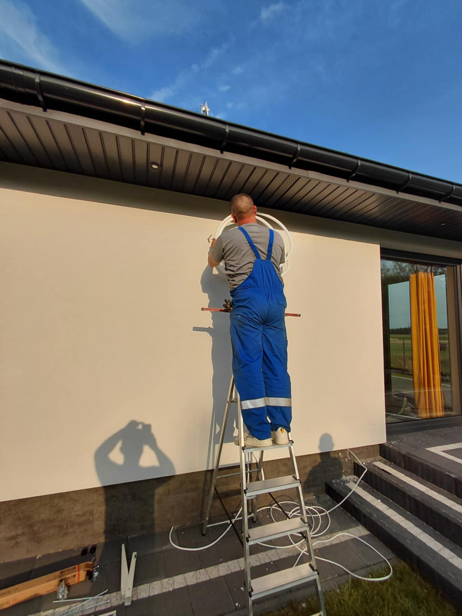 Instalator w niebieskim kombinezonie montuje białe rury na elewacji domu, stojąc na aluminiowej drabinie. Widoczne narzędzia i cień osoby robiącej zdjęcie.
