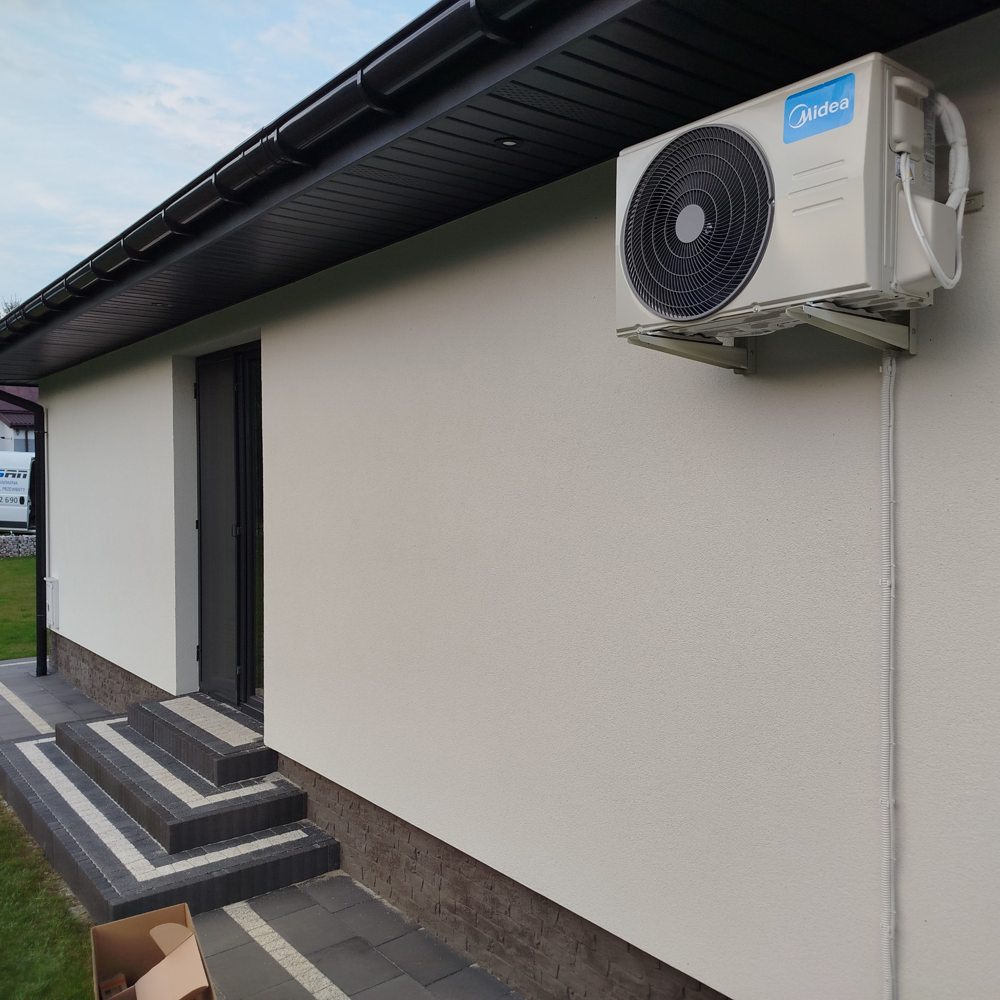 Zamontowana klimatyzacja Midea na elewacji domu z wykończonymi schodami wejściowymi. Widoczne kable w peszlu. Ujęcie z dołu, w tle fragment budynku.