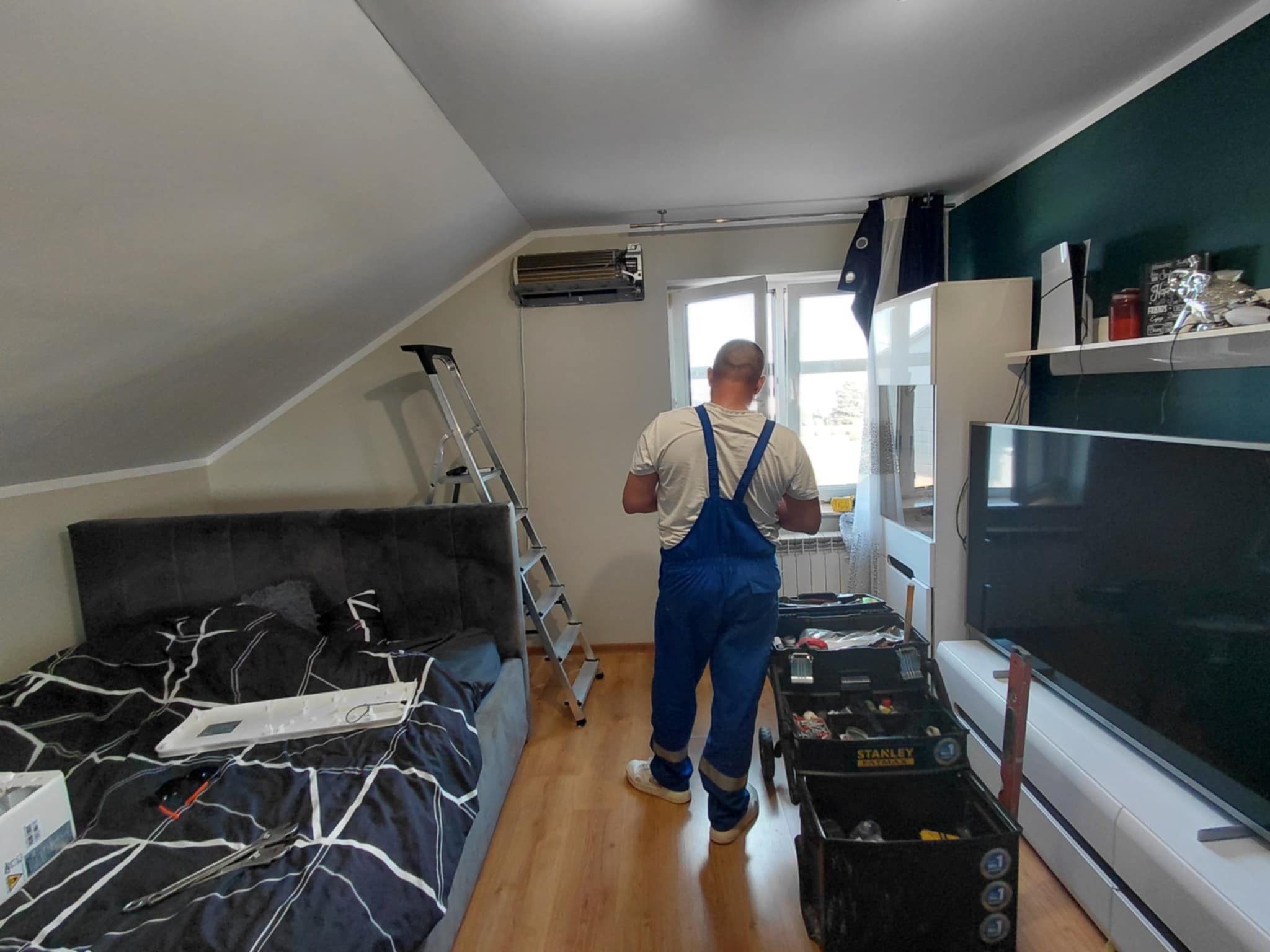Instalator w pokoju montuje klimatyzację. Widoczna drabina, narzędzia i zamontowana jednostka klimatyzacyjna na ścianie. Praca w trakcie wykonywania.