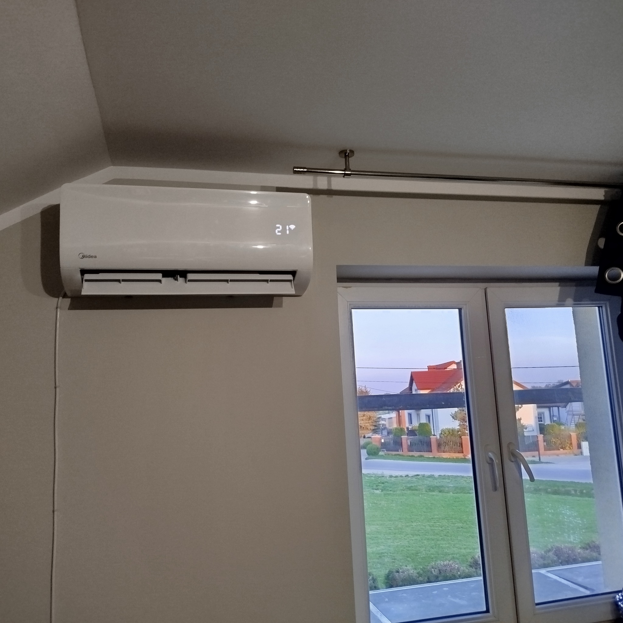 Zamontowany klimatyzator Midea na ścianie w pokoju z oknem. Widoczny wyświetlacz z temperaturą 21 stopni Celsjusza. Tło: budynki mieszkalne za oknem.