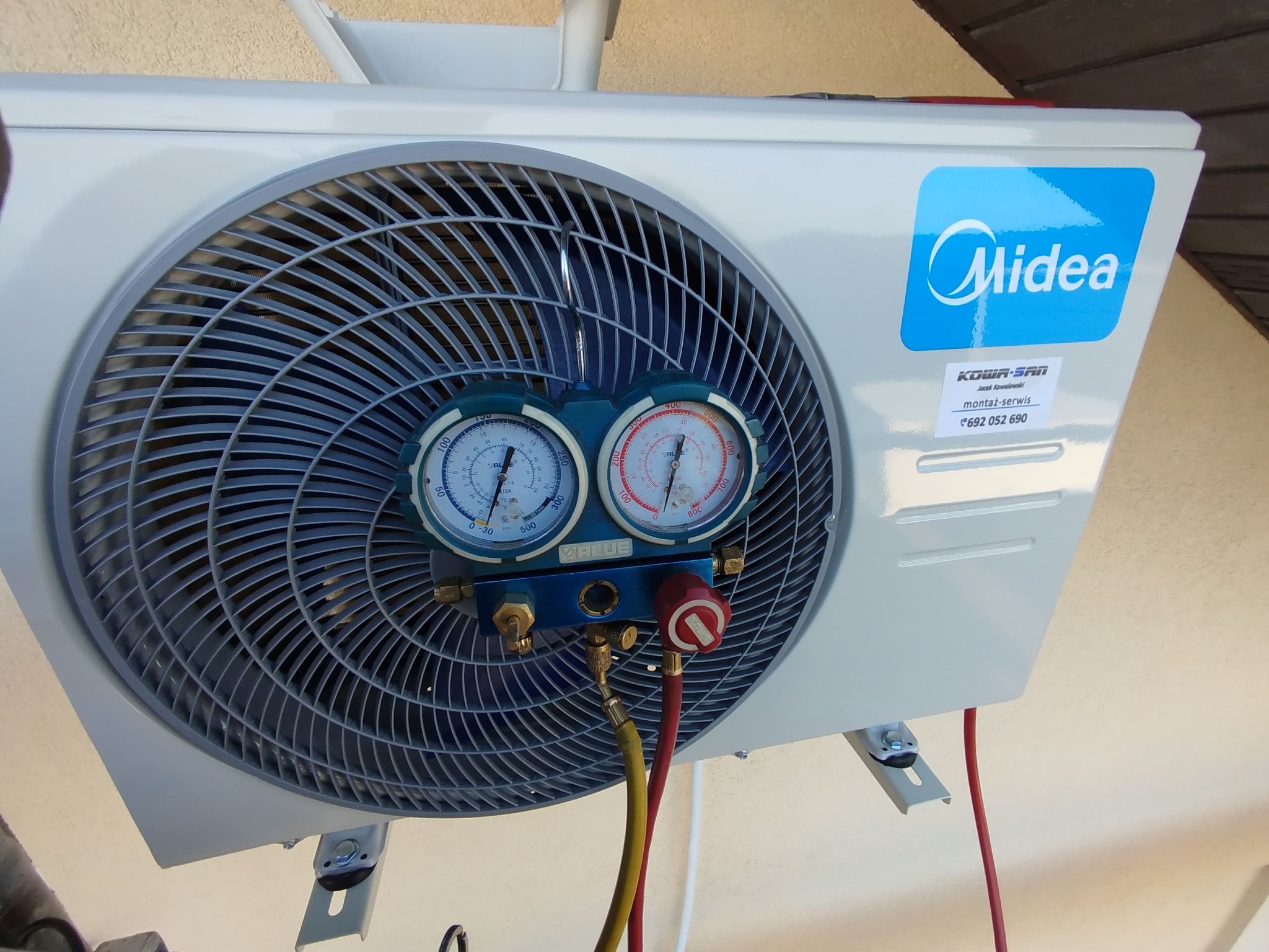 Biała jednostka zewnętrzna klimatyzacji Midea z podłączonymi manometrami serwisowymi podczas diagnostyki. Widoczne węże i skala ciśnienia.