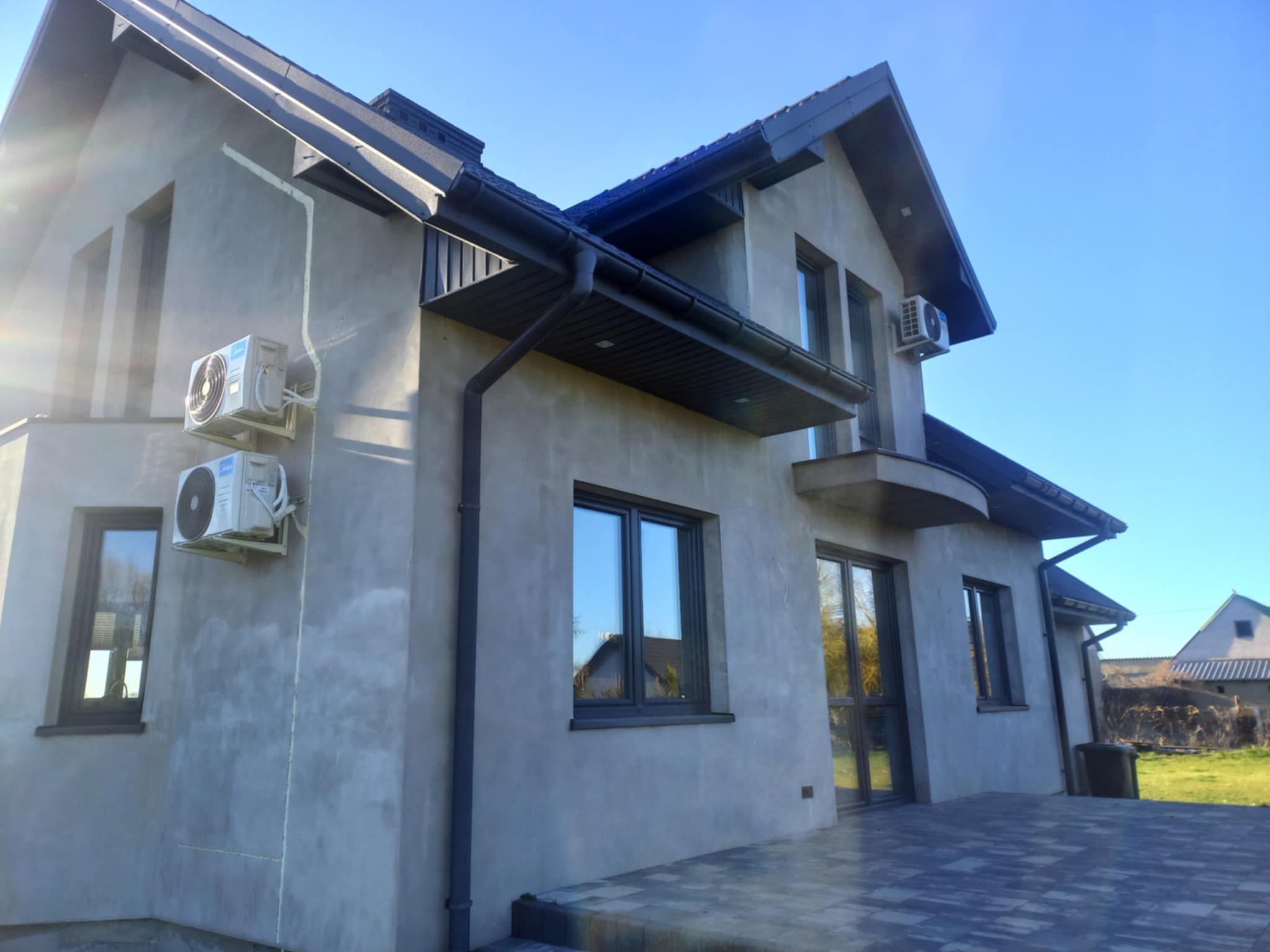 Szary dom jednorodzinny z zamontowanymi klimatyzatorami na elewacji i oknami z ciemną ramą. Widoczne rynny i fragment tarasu z kostki brukowej. Błękitne niebo w tle.