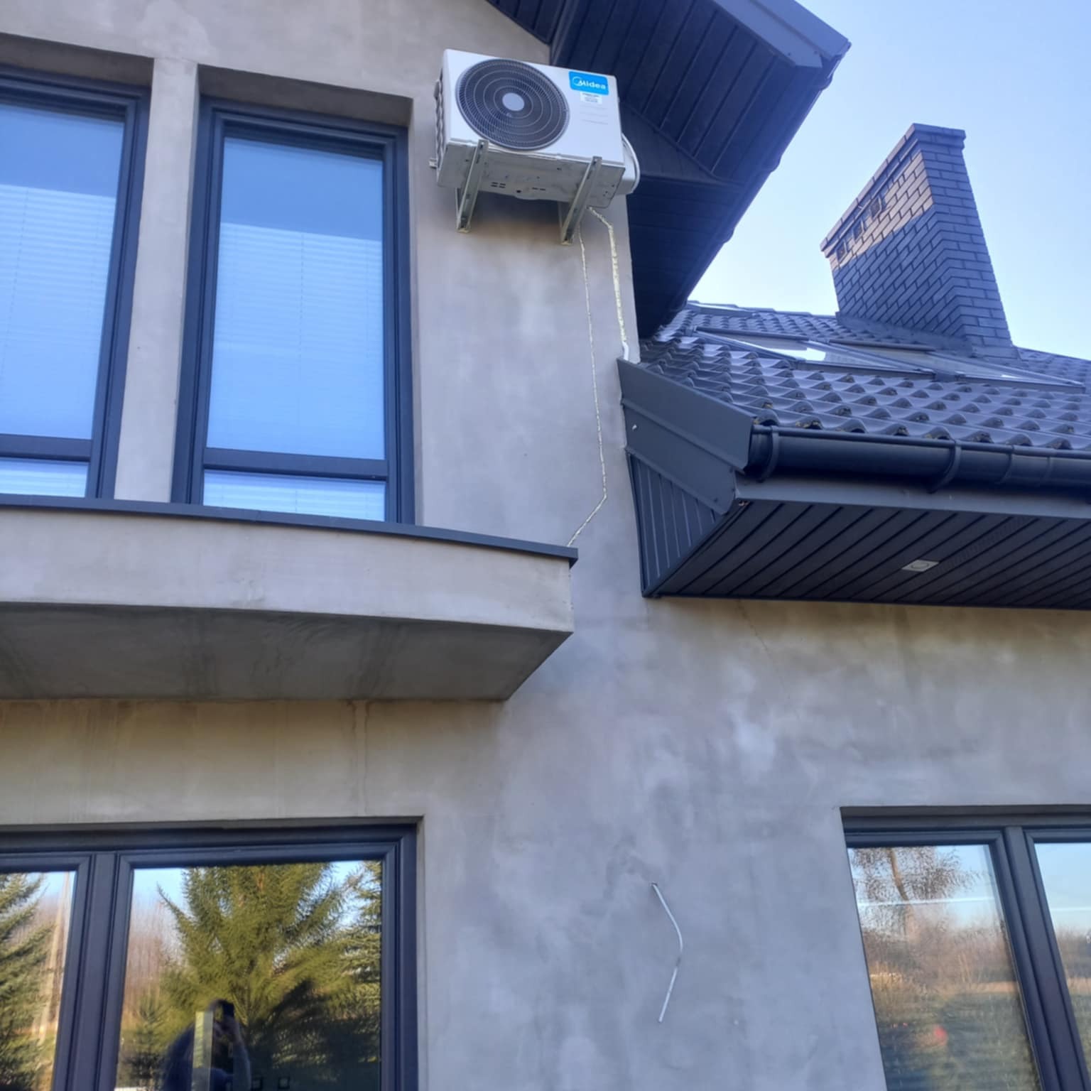 Biały klimatyzator Midea zamontowany na szarej elewacji domu z ciemnymi oknami i dachem. Widoczne przewody instalacji. Ujęcie z dołu, niebo w tle.