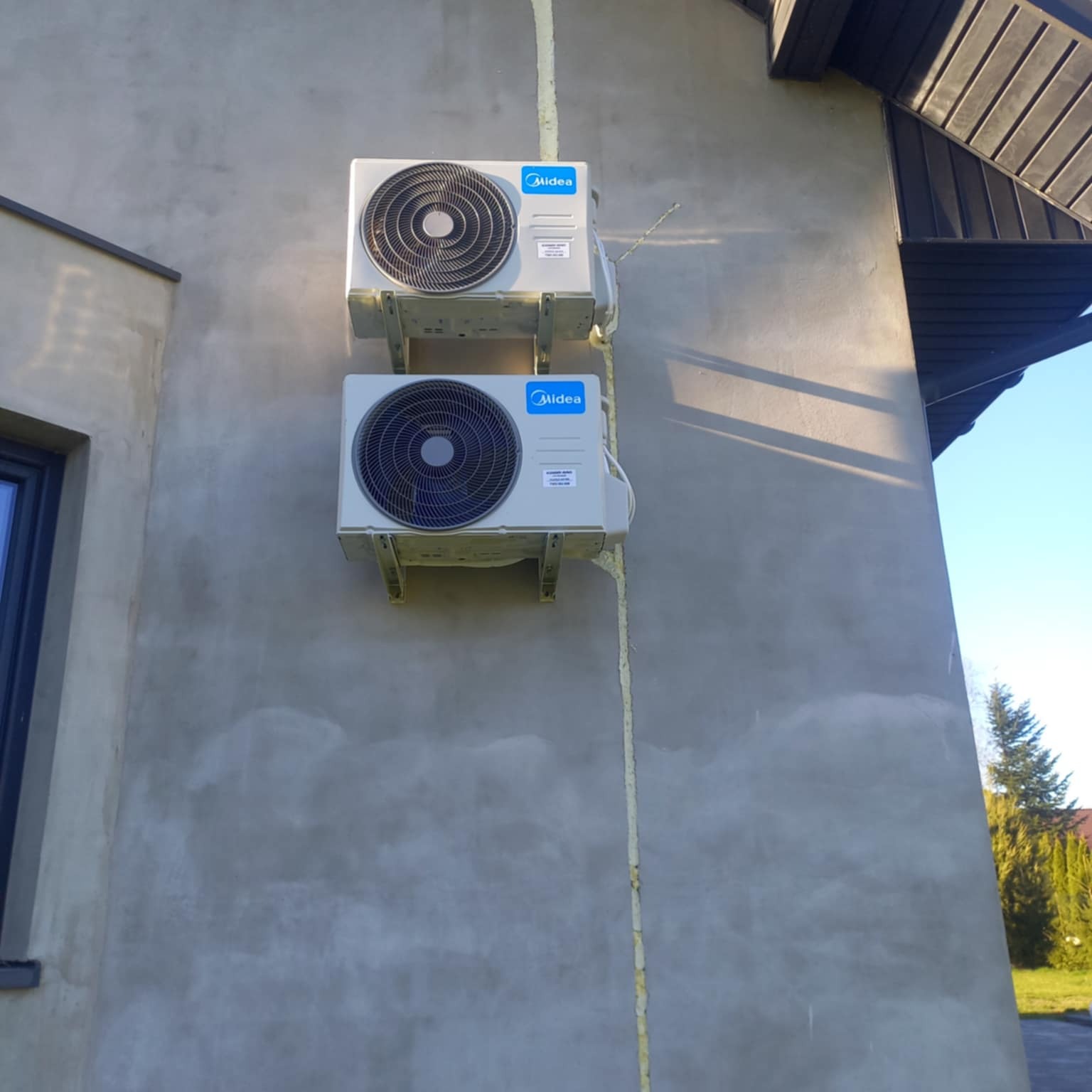Dwa klimatyzatory Midea zamontowane na szarej elewacji budynku jednorodzinnego. Widoczne elementy dachu i okno. Świeże uszczelnienie przy ścianie.