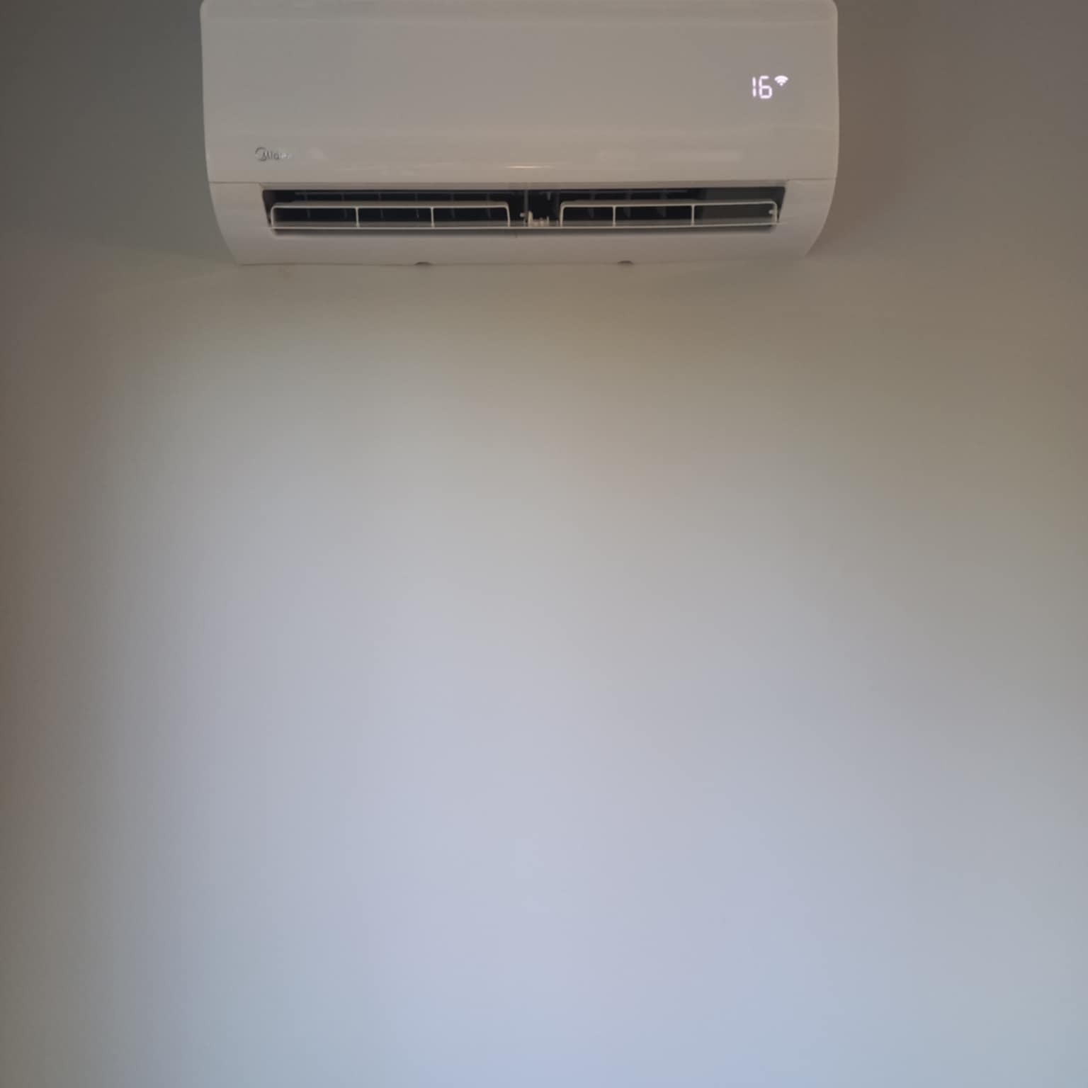 Biała klimatyzacja Midea na jasnej ścianie, ustawiona na 16 stopni. Widoczny wyświetlacz i kratka wentylacyjna. Minimalistyczny design.