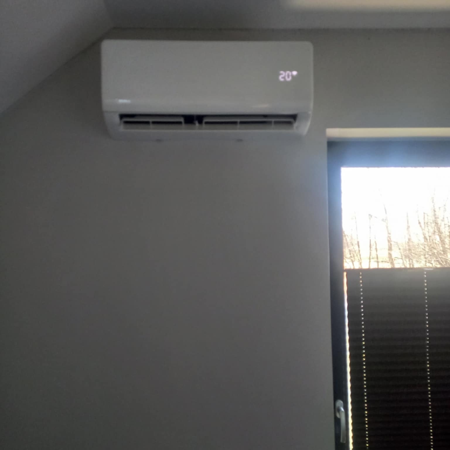 Biała klimatyzacja ścienna z wyświetlaczem 20 stopni Celsjusza w szarym pokoju, obok okna z żaluzjami. Widok z dołu.