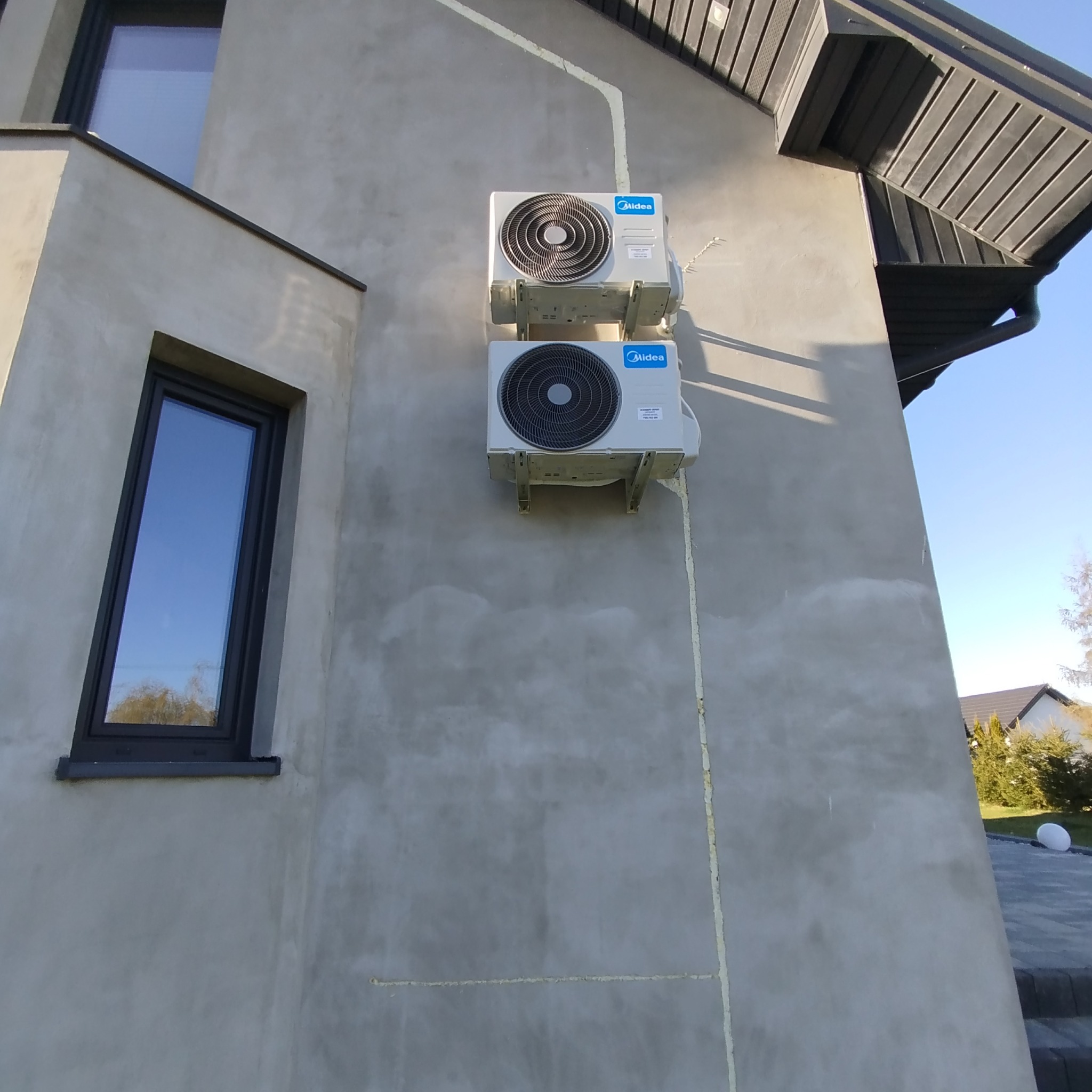Dwie jednostki zewnętrzne klimatyzacji Midea zamontowane na elewacji budynku z widocznymi przewodami i oknem. Widok z dołu na jasnym tle nieba.