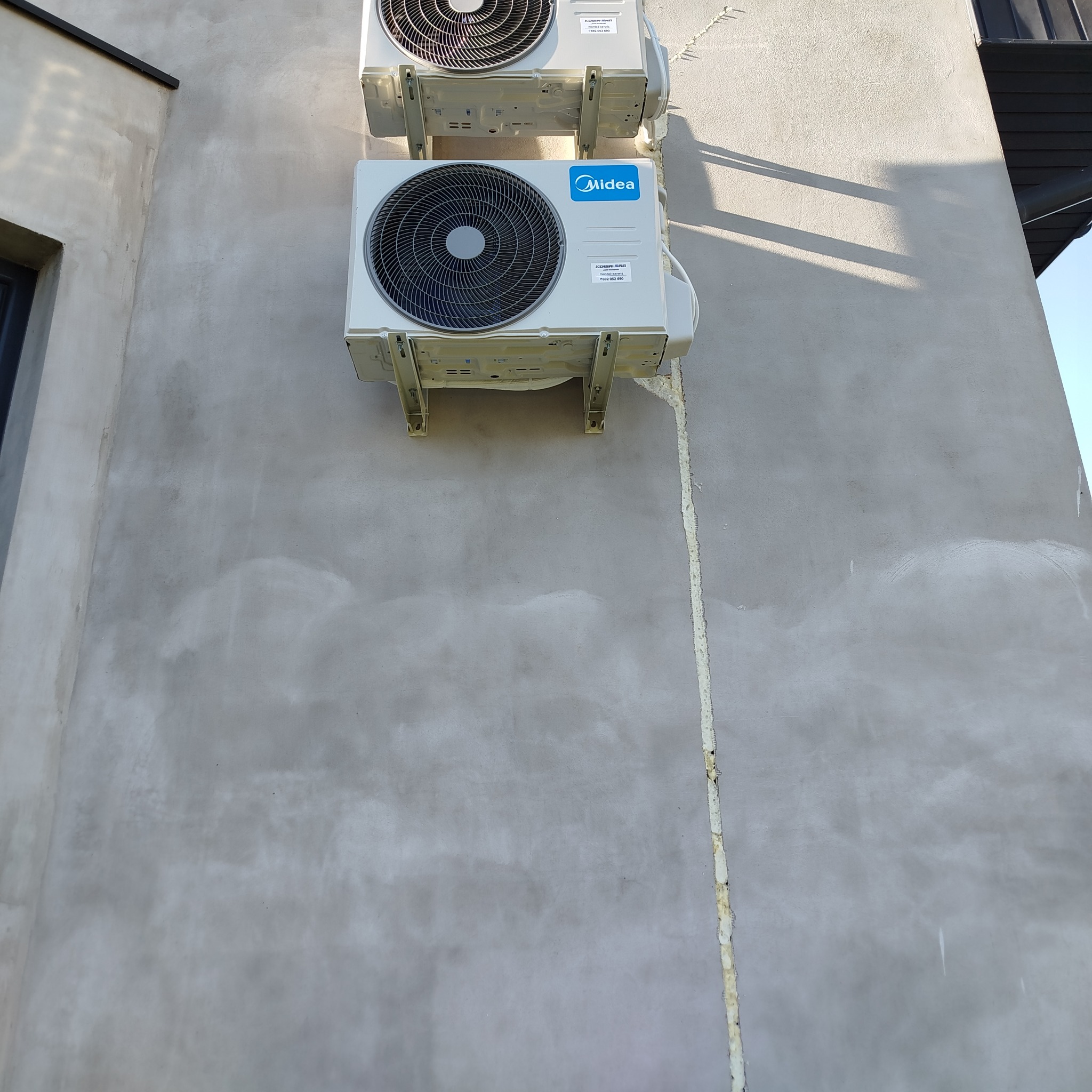 Dwa białe klimatyzatory Midea zamontowane na szarej, chropowatej ścianie budynku. Widoczne ślady pianki montażowej i cień rzucany przez elementy konstrukcyjne.