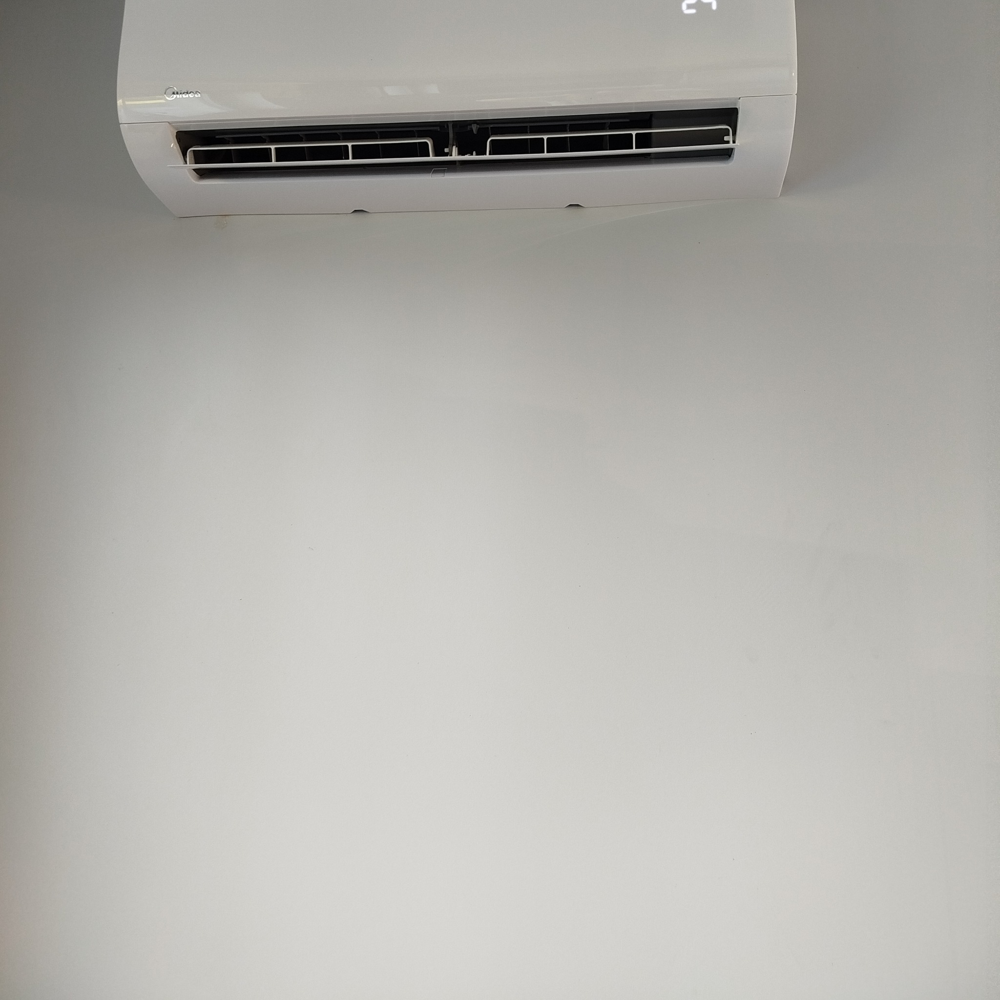 Biały klimatyzator Midea zamontowany na jasnej ścianie. Widoczny wyświetlacz temperatury i żaluzje wylotowe. Ujęcie od dołu, z dużą przestrzenią ściany pod urządzeniem.
