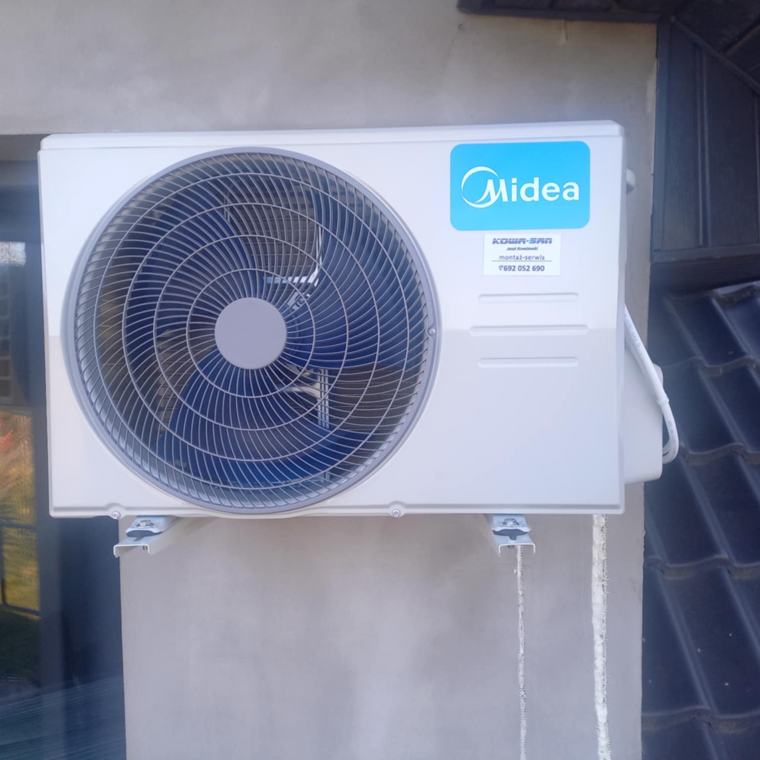 Zamontowana jednostka zewnętrzna klimatyzacji Midea na ścianie budynku, widoczne ślady montażu i uszczelnienia. Szare tło ściany kontrastuje z bielą urządzenia.