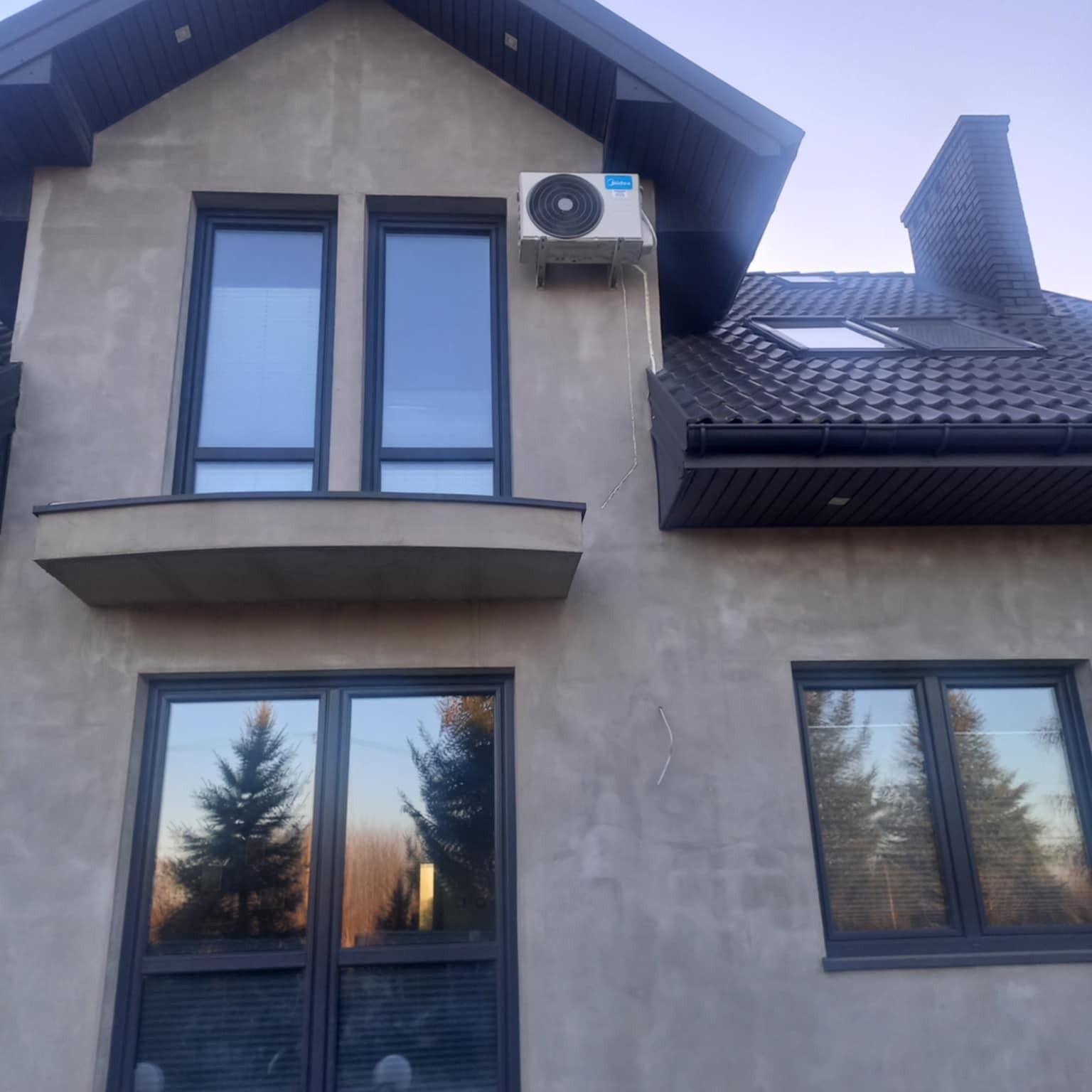 Szara elewacja domu z zamontowaną klimatyzacją Midea. Widoczne okna z ciemnymi ramami, dach z dachówki i komin. W oknach odbija się krajobraz z drzewami.