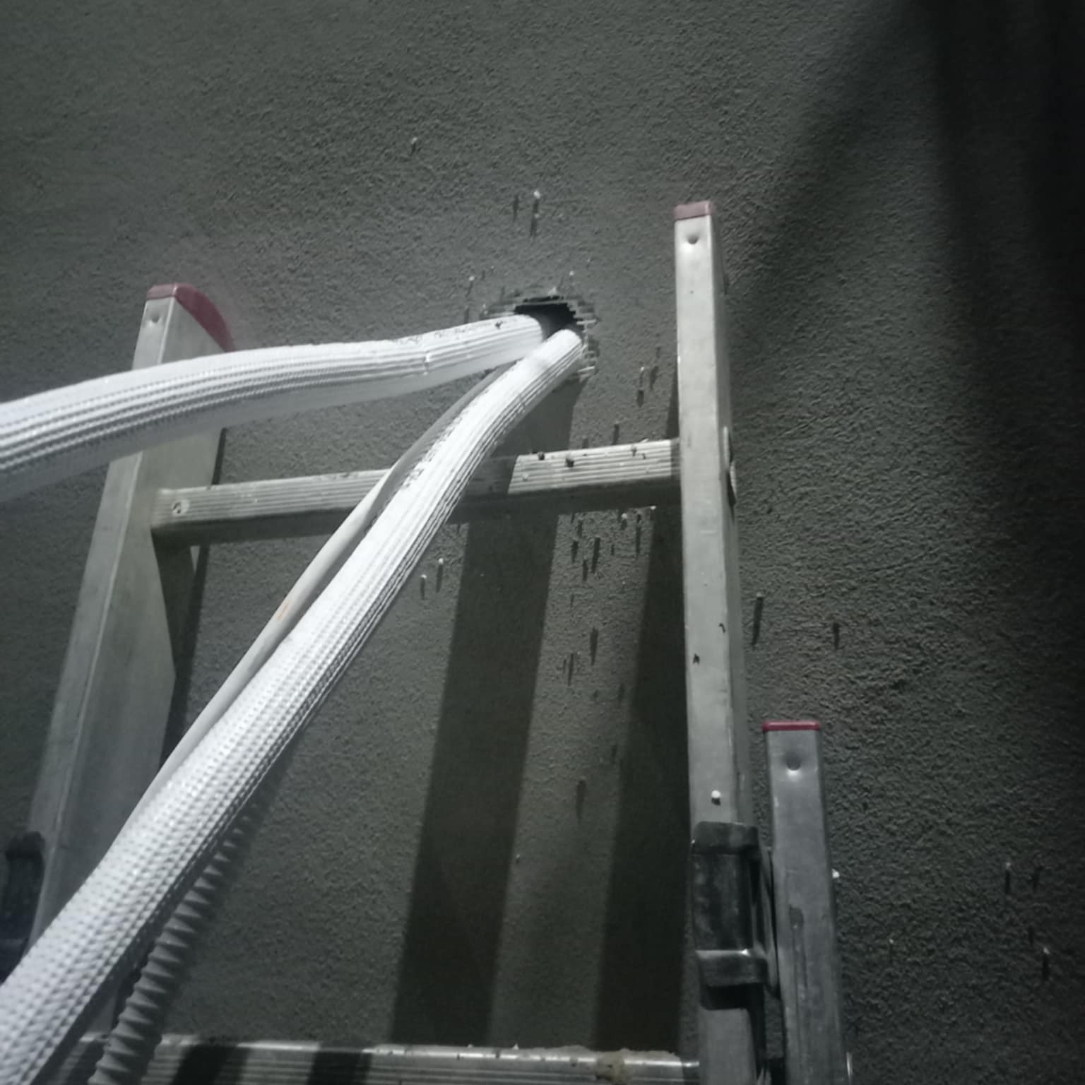 Białe, izolowane rury klimatyzacji wchodzące przez otwór w szarej ścianie, widoczne z drabiny aluminiowej. Instalacja w trakcie realizacji.