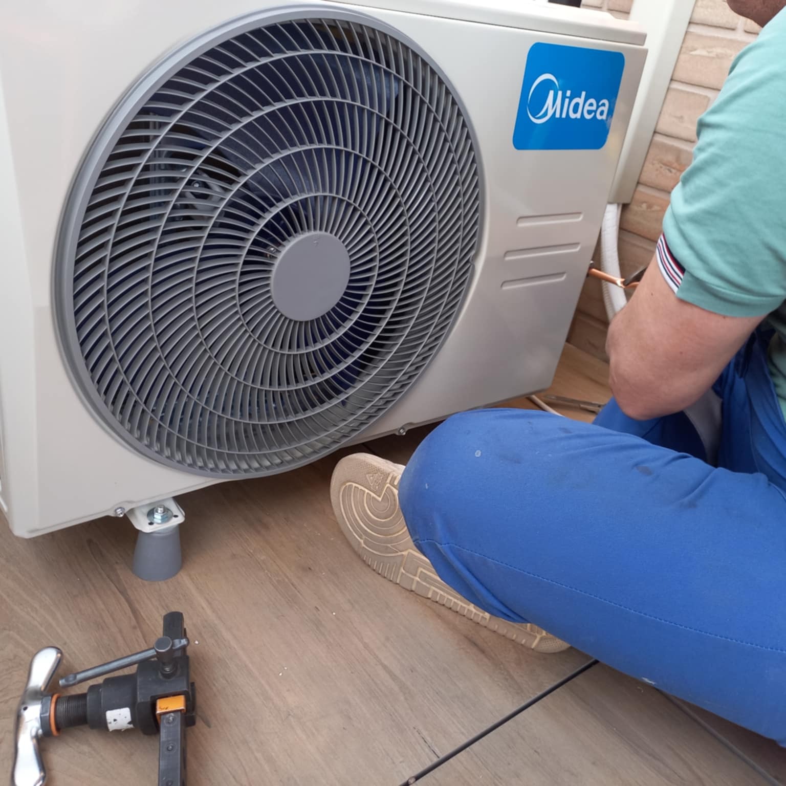 Montaż klimatyzacji Midea: technik w niebieskich spodniach łączy miedziane rurki przy zewnętrznej jednostce. Widoczne narzędzia i podstawa klimatyzatora.