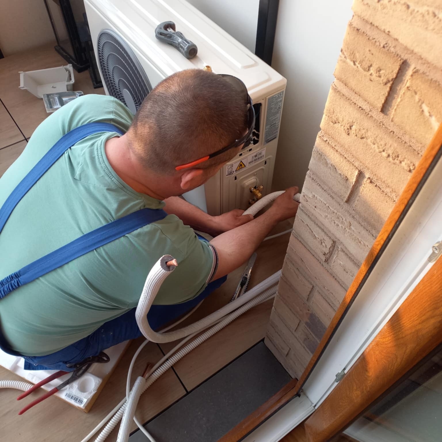 Instalator w trakcie montażu klimatyzacji w pomieszczeniu z ceglaną ścianą. Widoczne narzędzia i przewody. Kąt ujęcia z góry, oświetlenie dzienne.