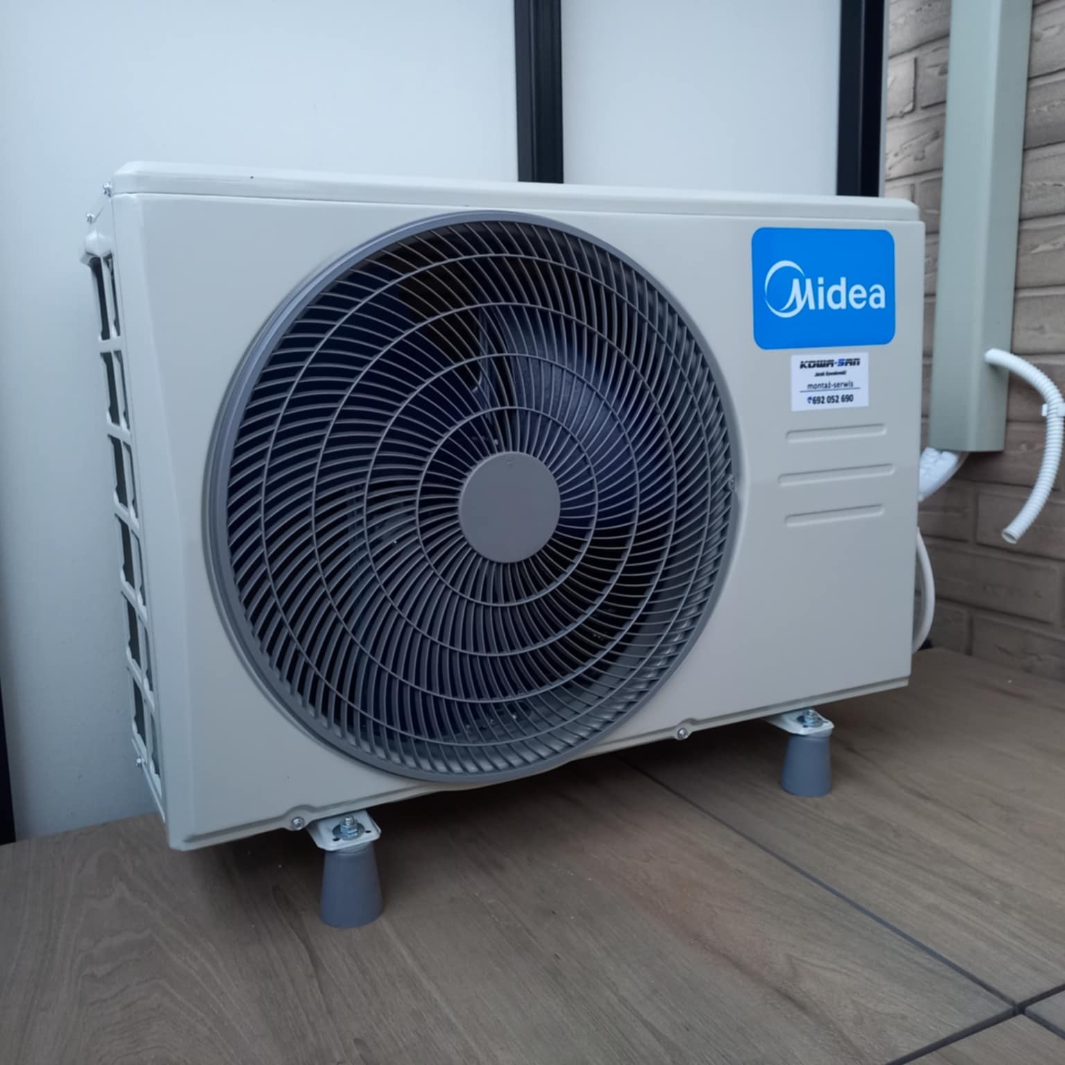 Zewnętrzna jednostka klimatyzacji Midea, zamontowana na podwyższeniu, z widocznym przewodem. Ściana z cegły w tle.