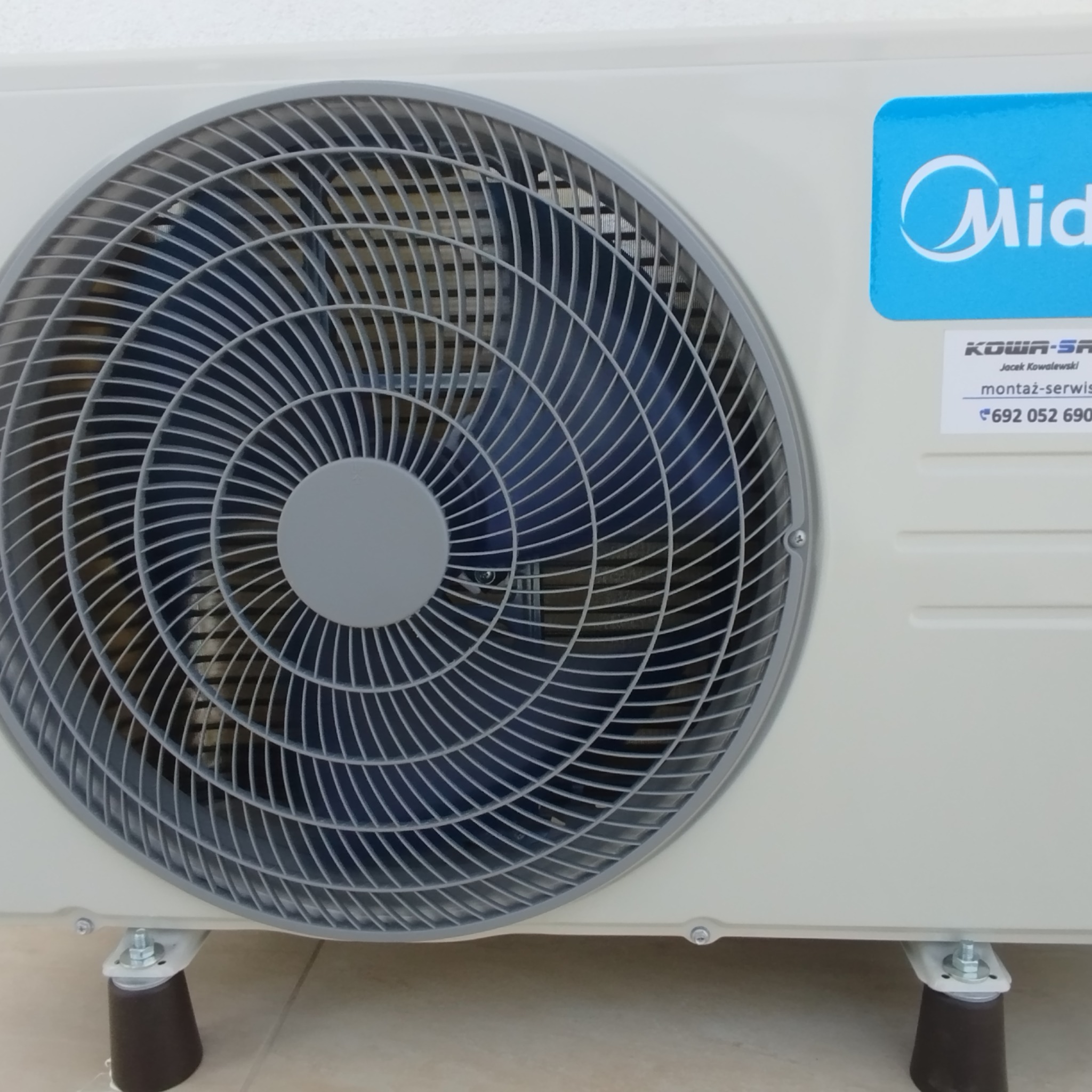 Zewnętrzna jednostka klimatyzacji Midea, z widocznym wentylatorem i naklejką z danymi kontaktowymi montera na gładkiej, białej ścianie. Widoczne gumowe nóżki.