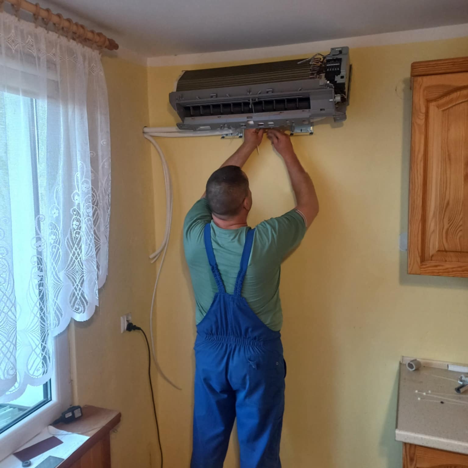 Mężczyzna w niebieskim kombinezonie montuje klimatyzator na żółtej ścianie, widoczne okno z firanką i szafka kuchenna. Praca w trakcie instalacji.