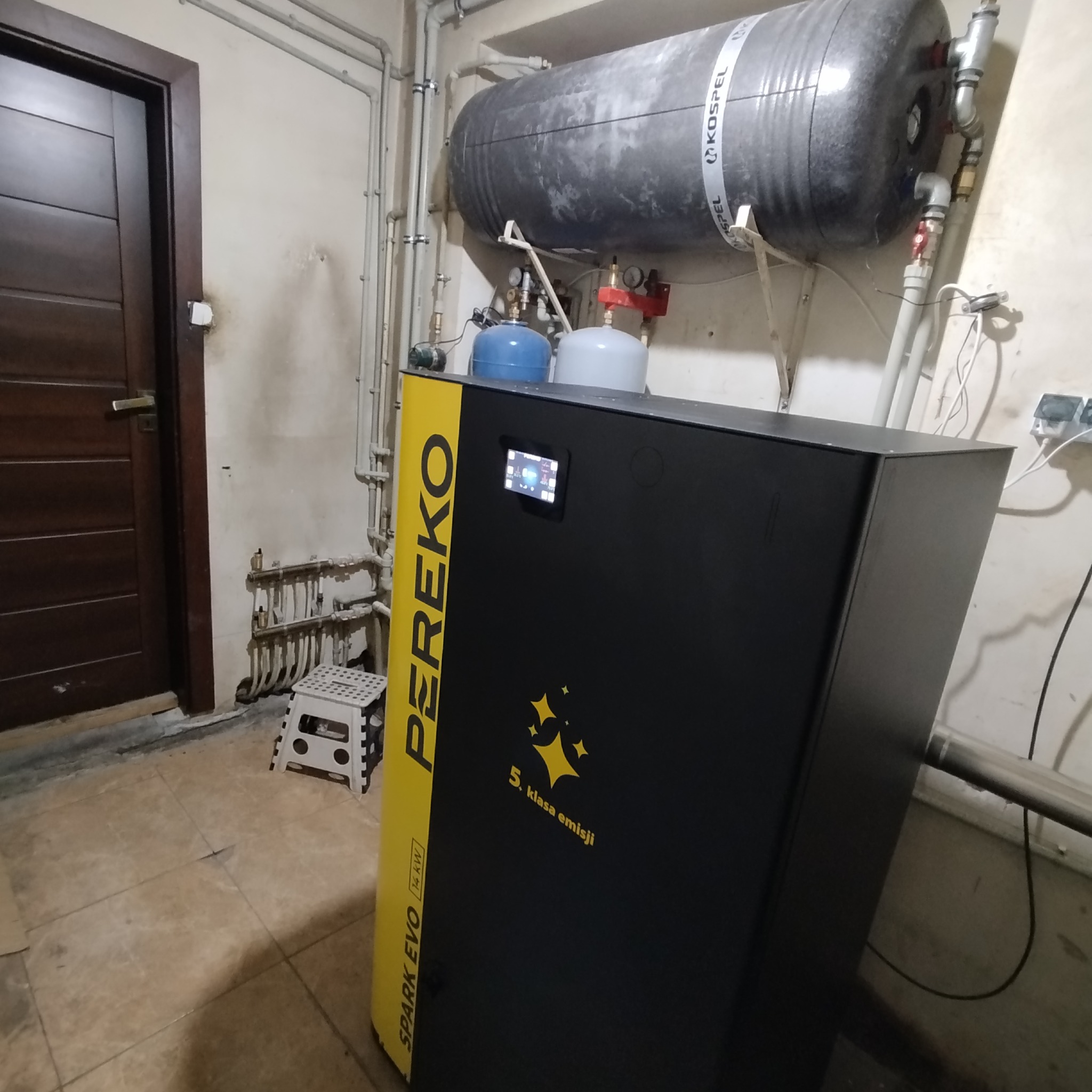 Piec Pereko Spark Evo 14kW z zbiornikiem Kospel, widoczny w kotłowni. Żółto-czarna obudowa pieca z panelem sterowania. Rury instalacji grzewczej w tle.