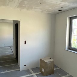 Grupa MFC sp. z o.o. - Wnętrze w trakcie remontu: sufit z otworami na oświetlenie, gładzie na ścianach, przygotowane podłoże. Widoczna puszka elektryczna i karton. Jasne, naturalne światło.