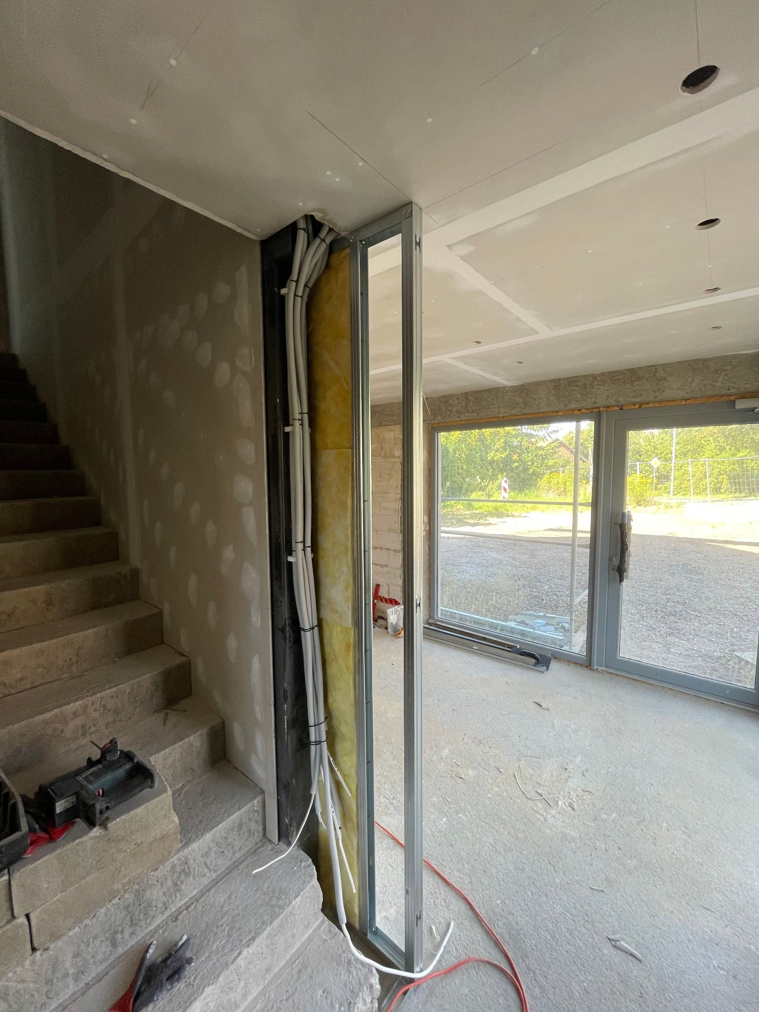 Ściana w trakcie budowy z widocznymi przewodami elektrycznymi prowadzonymi wewnątrz konstrukcji szkieletowej, z izolacją z wełny mineralnej. W tle schody i okno.
