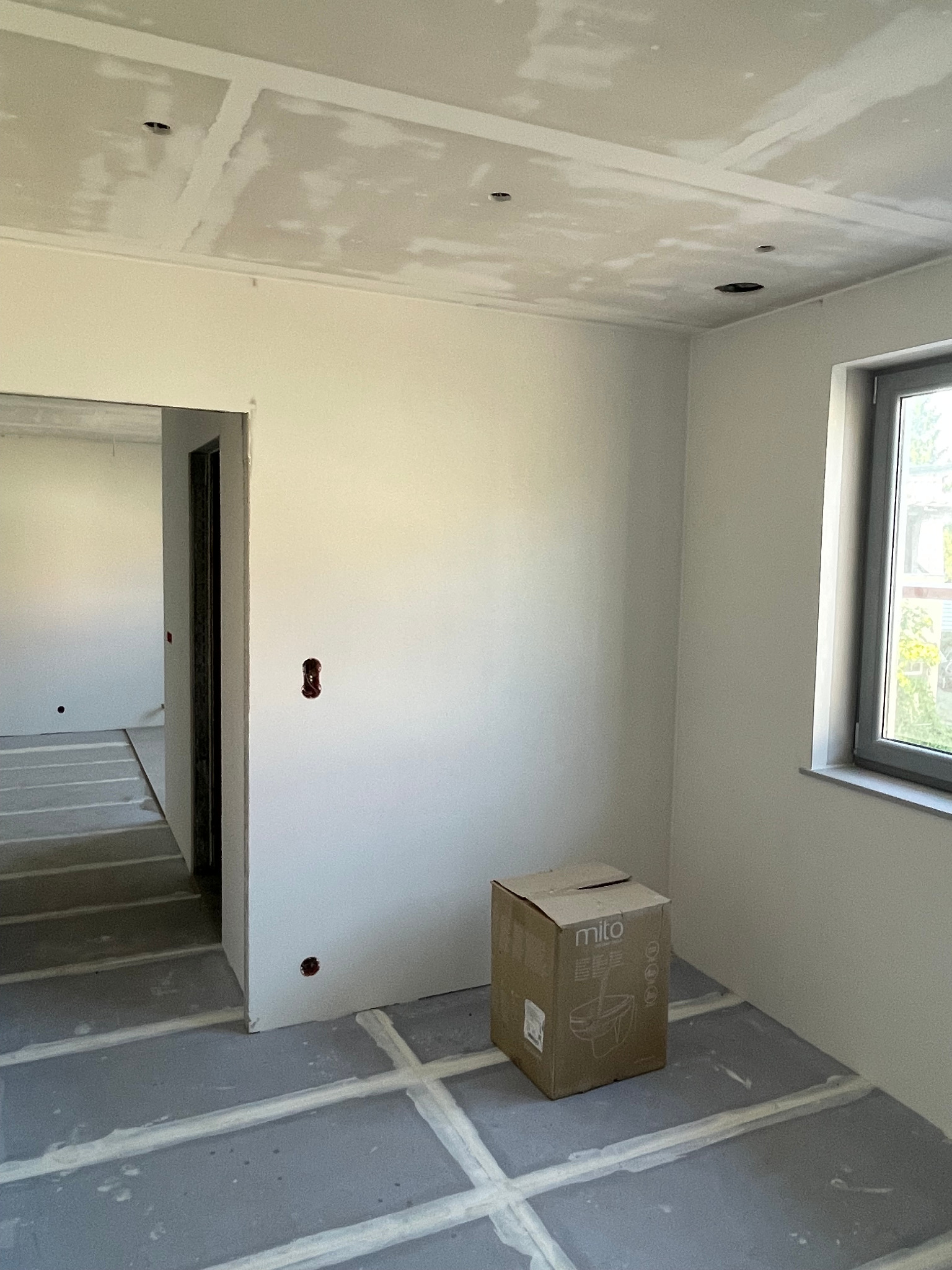 Wnętrze w trakcie remontu: sufit z otworami na oświetlenie, gładzie na ścianach, przygotowane podłoże. Widoczna puszka elektryczna i karton. Jasne, naturalne światło.