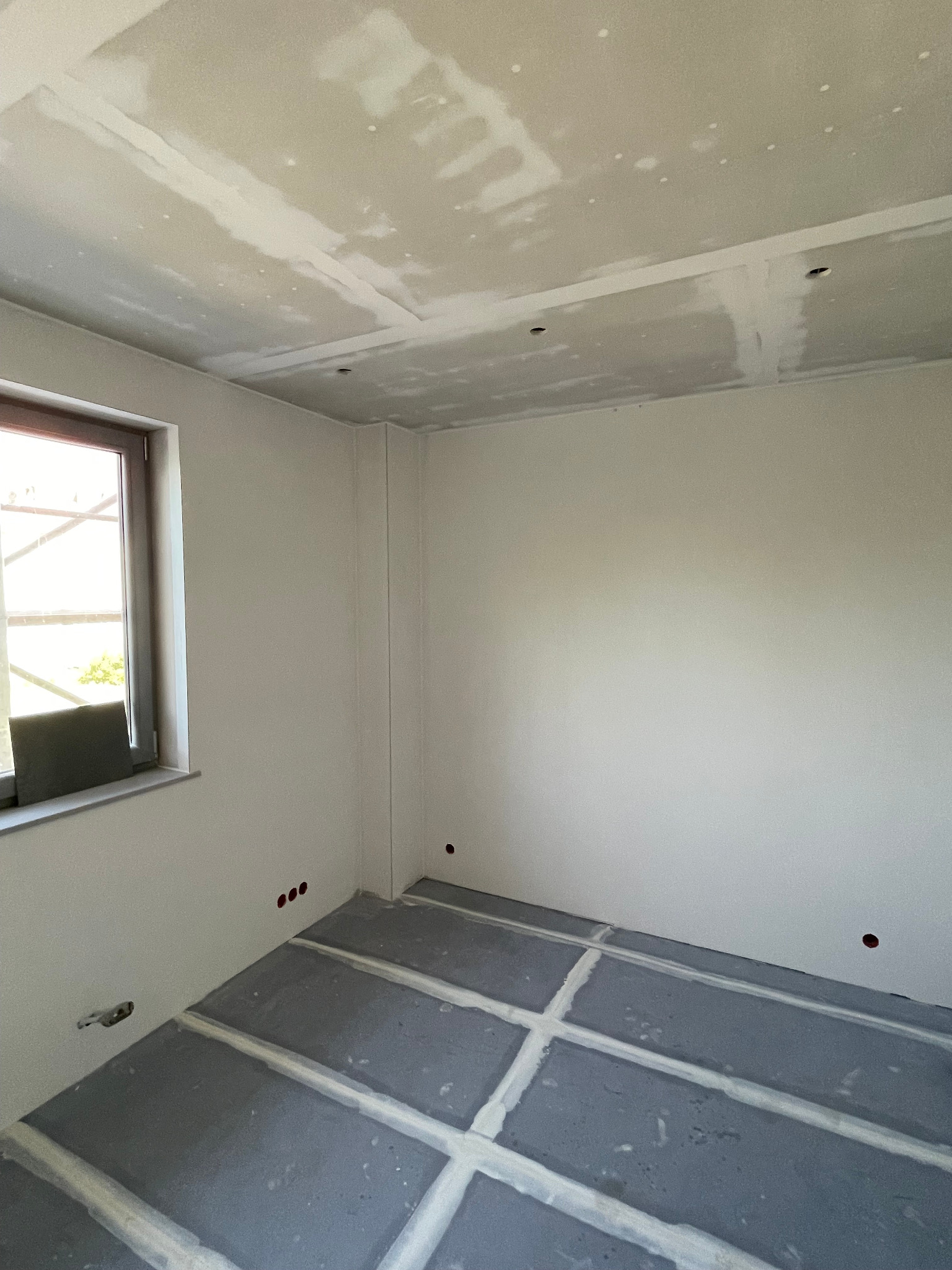 Wnętrze w trakcie prac wykończeniowych: ściany i sufit po szpachlowaniu, podłoga z widocznymi liniami pod przyszłą instalację. Widoczne puszki elektryczne.