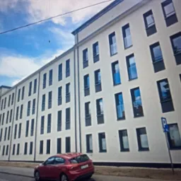 Biały budynek wielorodzinny z rzędami okien z czarnymi ramami, odbijających niebo, z zaparkowanym czerwonym autem i znakiem parkingu.