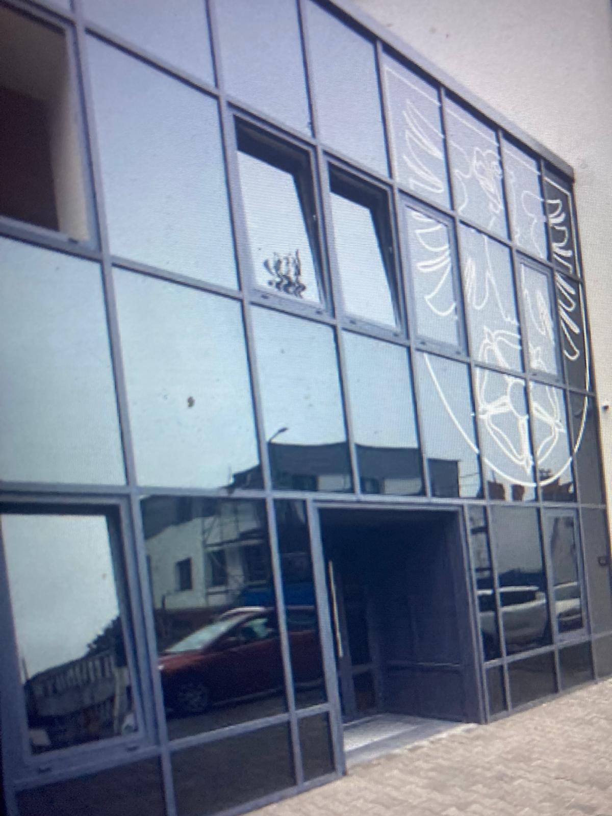 Fasada budynku z dużymi, szklanymi oknami w ciemnych ramach, z białym, abstrakcyjnym wzorem na części okien. W oknach odbija się otoczenie i samochód.