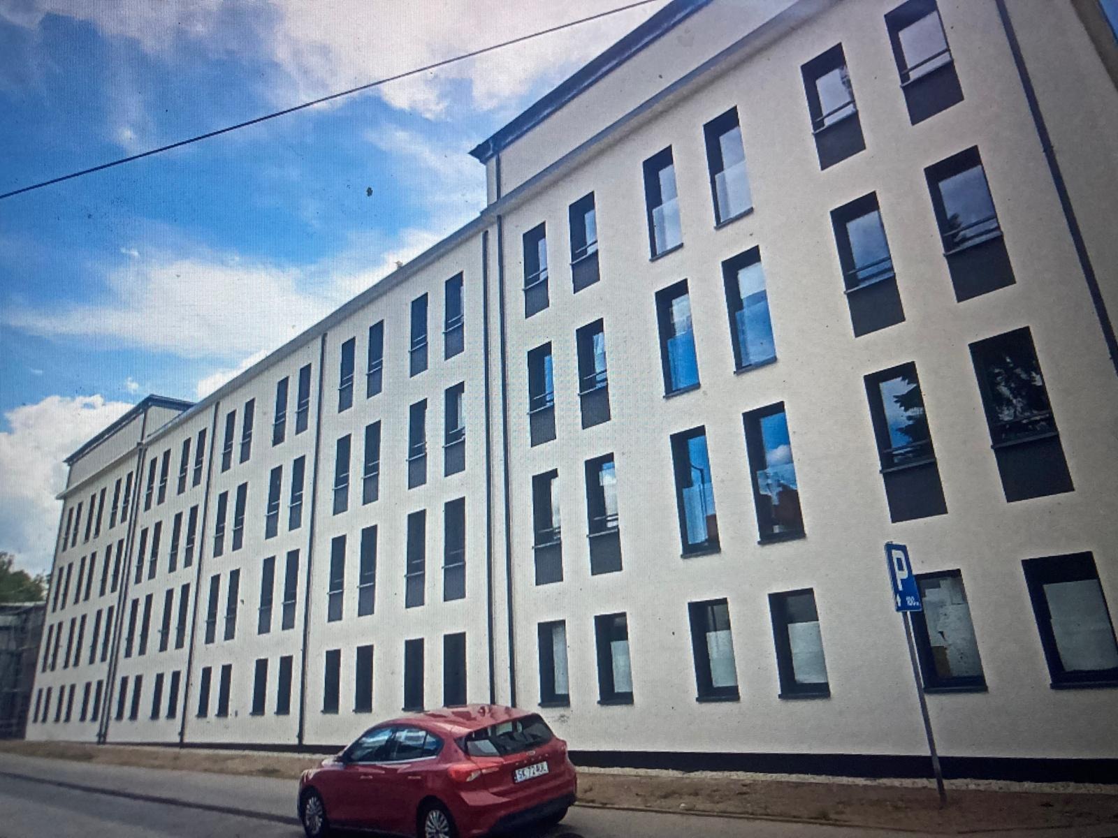 Biały budynek wielorodzinny z rzędami okien z czarnymi ramami, odbijających niebo, z zaparkowanym czerwonym autem i znakiem parkingu.