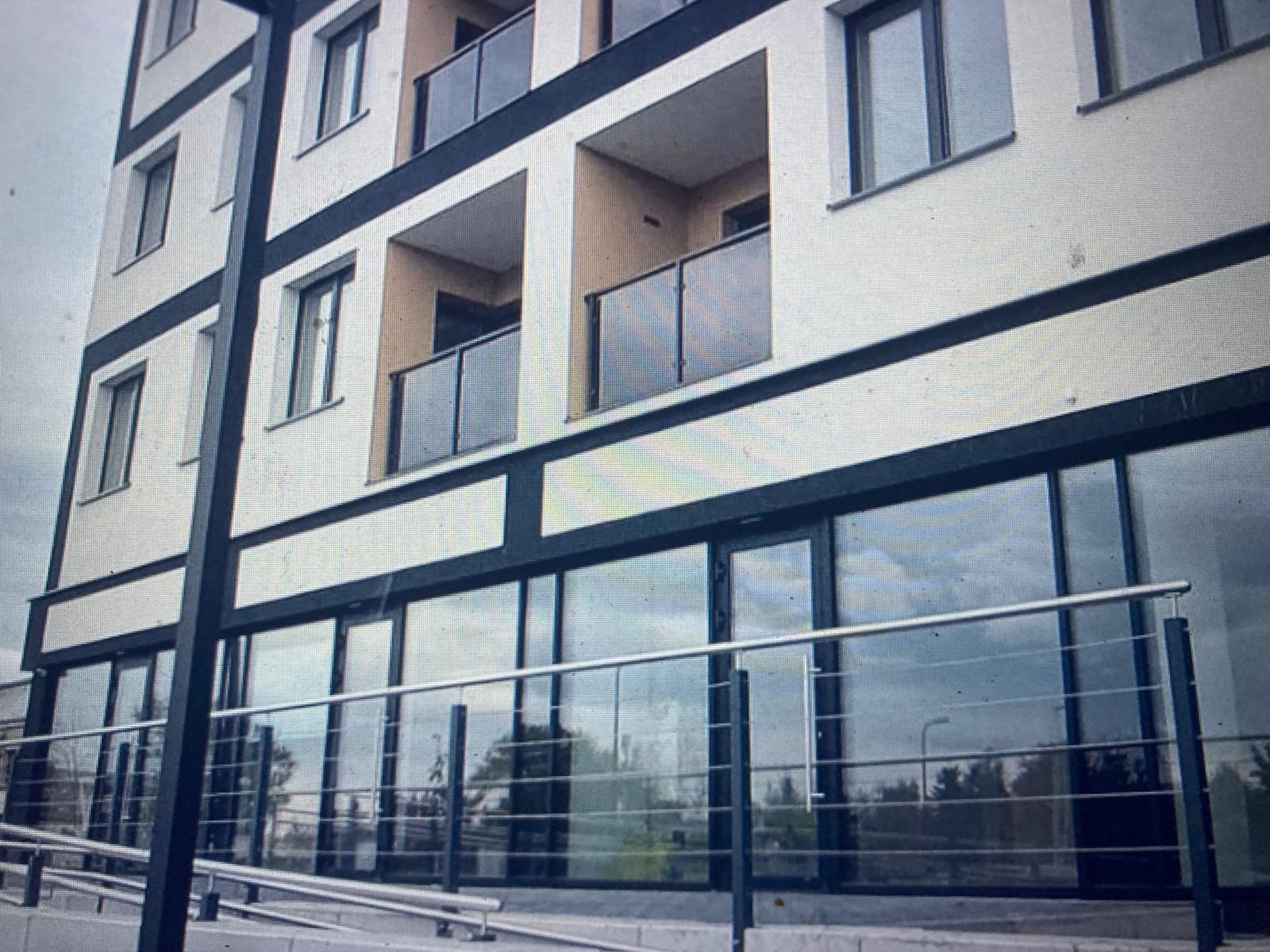 Nowoczesny budynek z balkonami i dużymi przeszkleniami na parterze. Widoczne barierki balkonowe ze szkła i metalu. Elewacja w jasnych kolorach z ciemnymi akcentami.