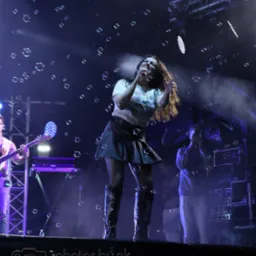 Wokalistka na scenie, otoczona bańkami mydlanymi, w świetle reflektorów. W tle widoczny gitarzysta i sprzęt muzyczny. Dynamiczne ujęcie z koncertu.