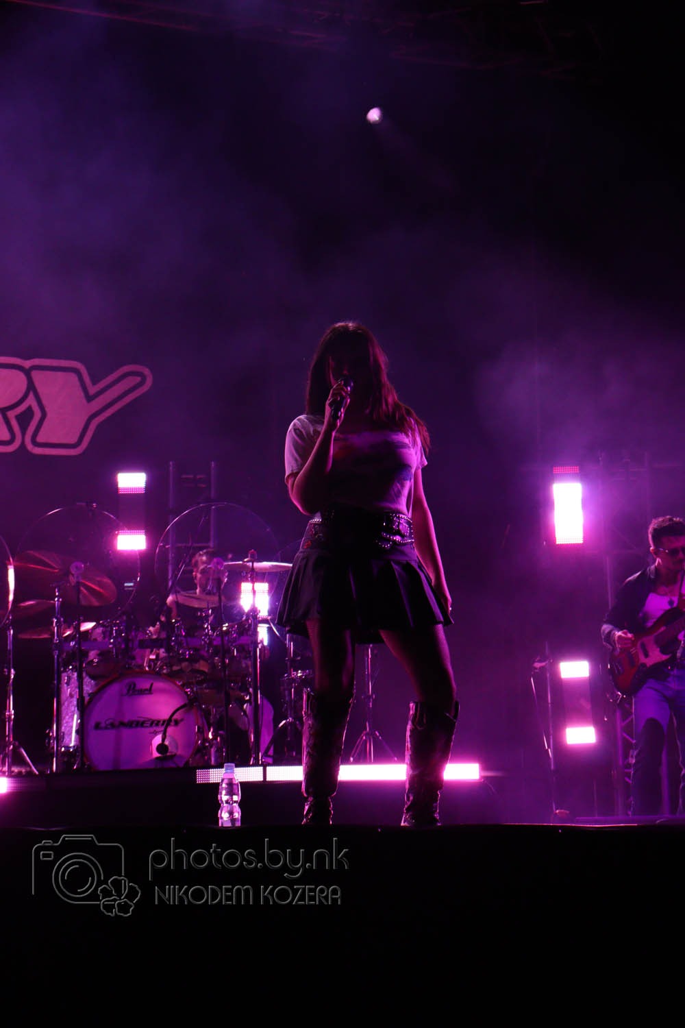 Piosenkarka z mikrofonem na scenie w fioletowym oświetleniu, za nią perkusja i gitarzysta. Na pierwszym planie butelka wody i logo fotografa.