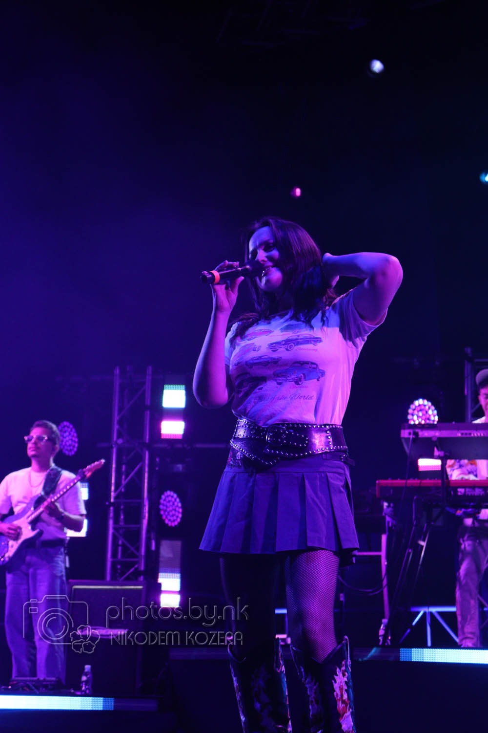 Wokalistka z mikrofonem na scenie w fioletowym oświetleniu, z zespołem muzycznym w tle. Uwagę przykuwają kowbojskie buty i szeroki pas z ćwiekami.