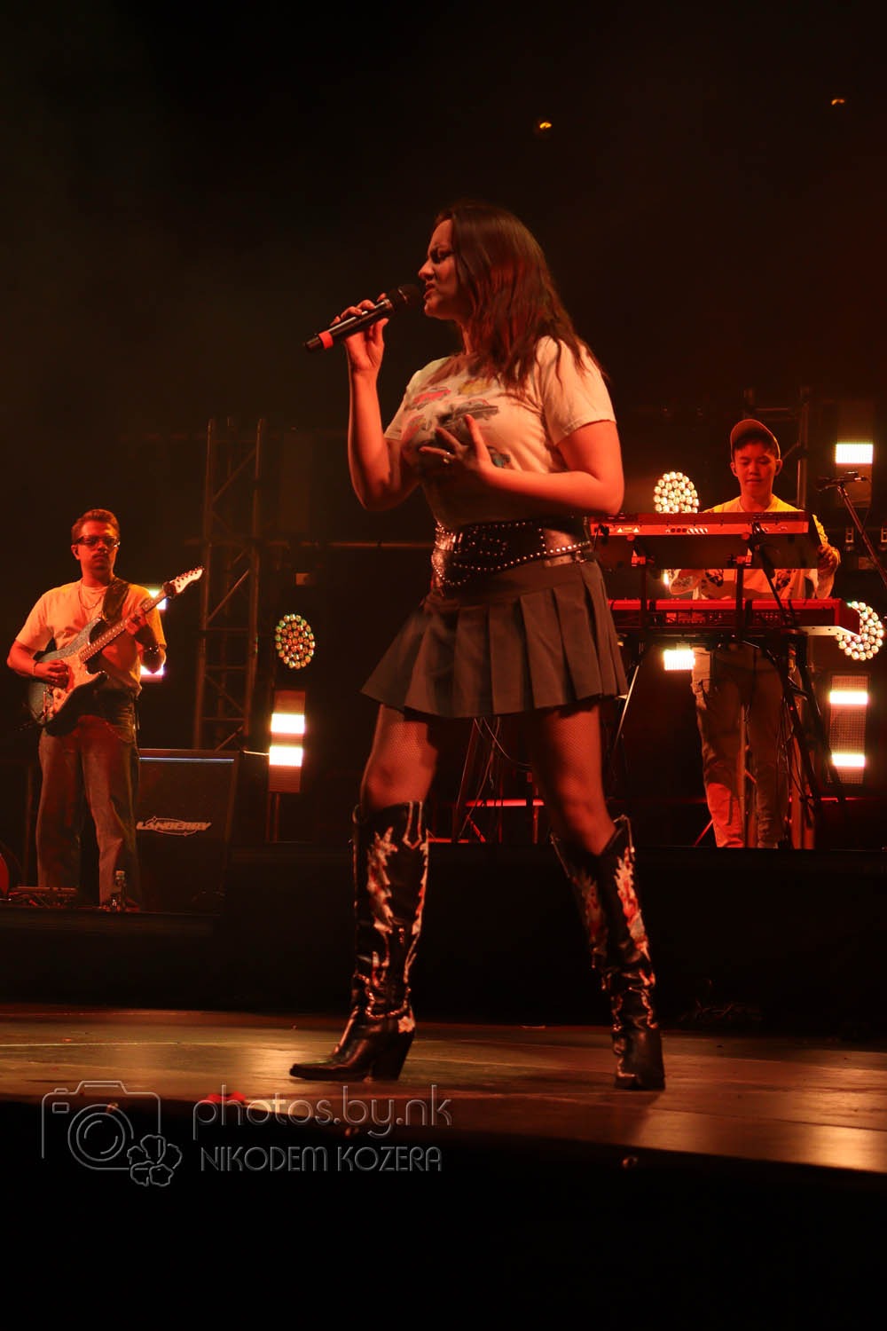 Piosenkarka z mikrofonem w dłoni, w otoczeniu muzyków z gitarą i keyboardem na scenie, w blasku reflektorów podczas koncertu.