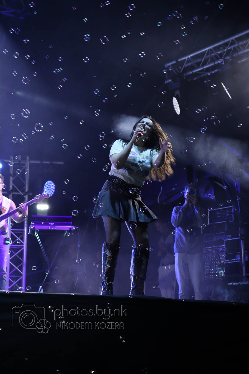 Wokalistka na scenie, otoczona bańkami mydlanymi, w świetle reflektorów. W tle widoczny gitarzysta i sprzęt muzyczny. Dynamiczne ujęcie z koncertu.
