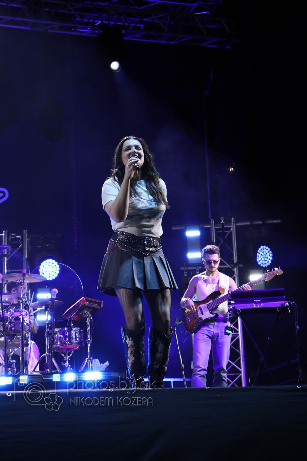 Wokalistka śpiewa na scenie podczas koncertu, w tle perkusja i basista. Scena oświetlona niebieskim światłem.