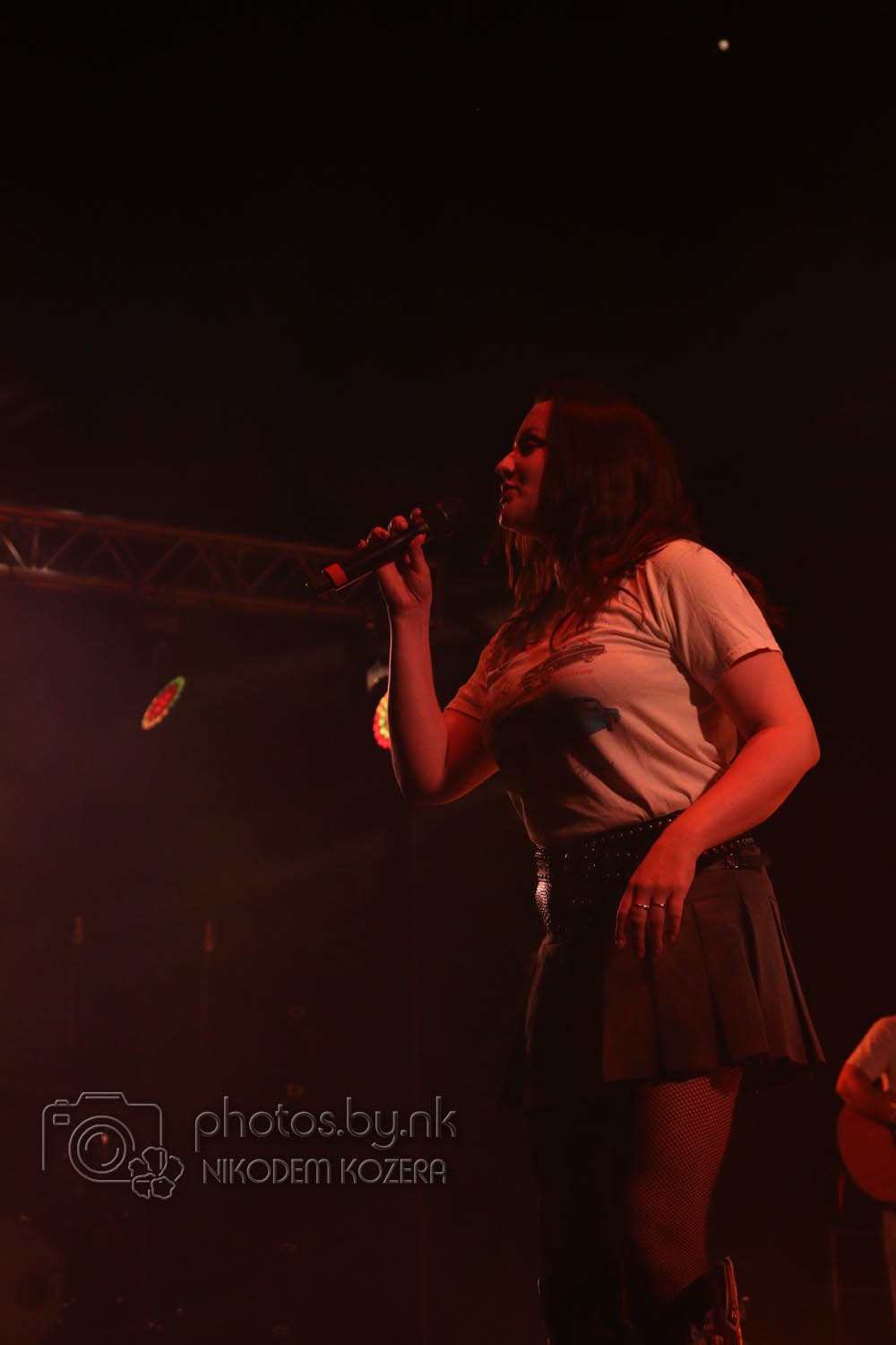 Wokalistka z mikrofonem na scenie w czerwonym oświetleniu. Artystka w koszulce i spódnicy, z ciemnymi włosami, śpiewa podczas koncertu. Widoczne elementy scenografii.