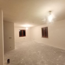 Tomasz M - Wnętrze świeżo wykończonego pokoju z dwoma oknami w drewnianych ramach. Ściany i sufit są w jasnym odcieniu, podłoga szara. Widoczne punkty oświetleniowe.