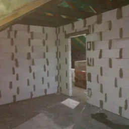 Wnętrze domu w trakcie budowy: białe ściany z betonu komórkowego z widocznymi spoinami, drewniana konstrukcja dachu, surowa podłoga, otwór drzwiowy.