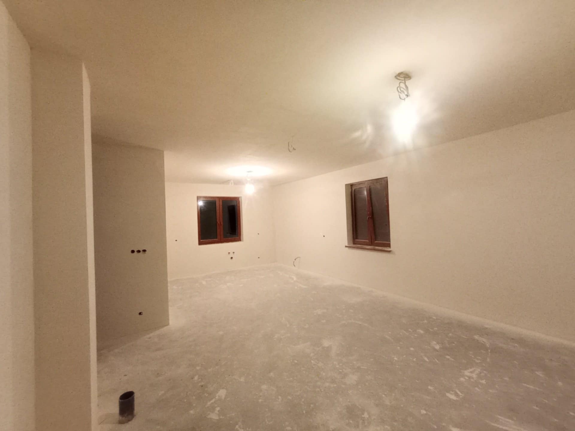 Wnętrze świeżo wykończonego pokoju z dwoma oknami w drewnianych ramach. Ściany i sufit są w jasnym odcieniu, podłoga szara. Widoczne punkty oświetleniowe.