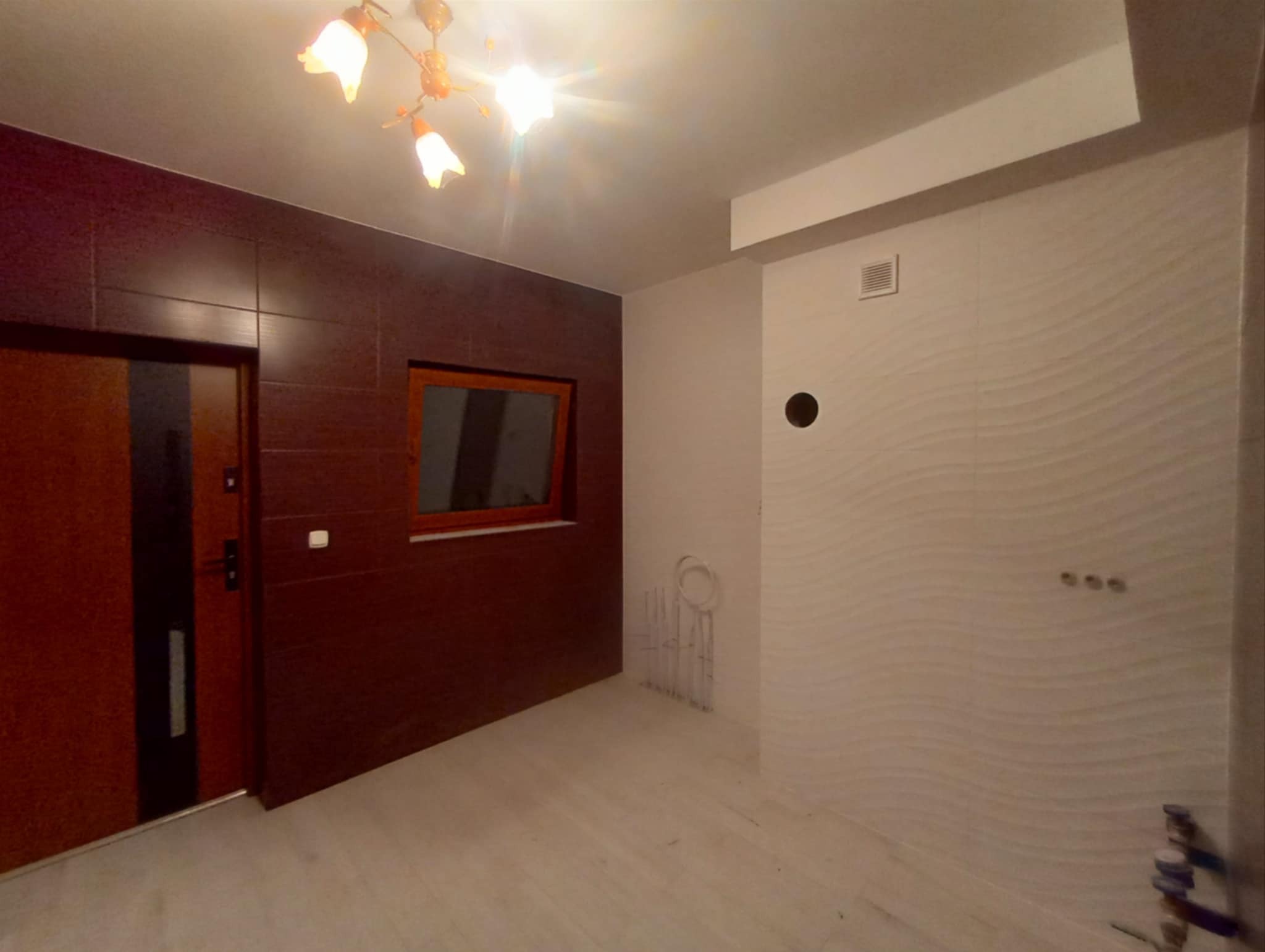 Wnętrze po remoncie: brązowe płytki na ścianie z drzwiami i oknem, biała ściana z dekoracyjną fakturą. Widoczne rury instalacji i designerska lampa sufitowa.