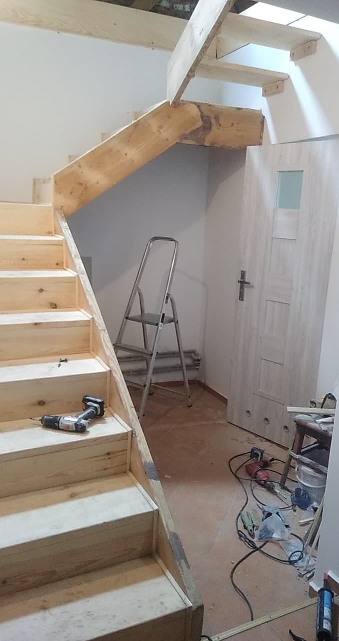 Montaż drewnianych schodów wewnątrz budynku. Widoczne narzędzia: wkrętarka, drabina. Surowe drewno, jasne wnętrze. Etap budowy schodów.