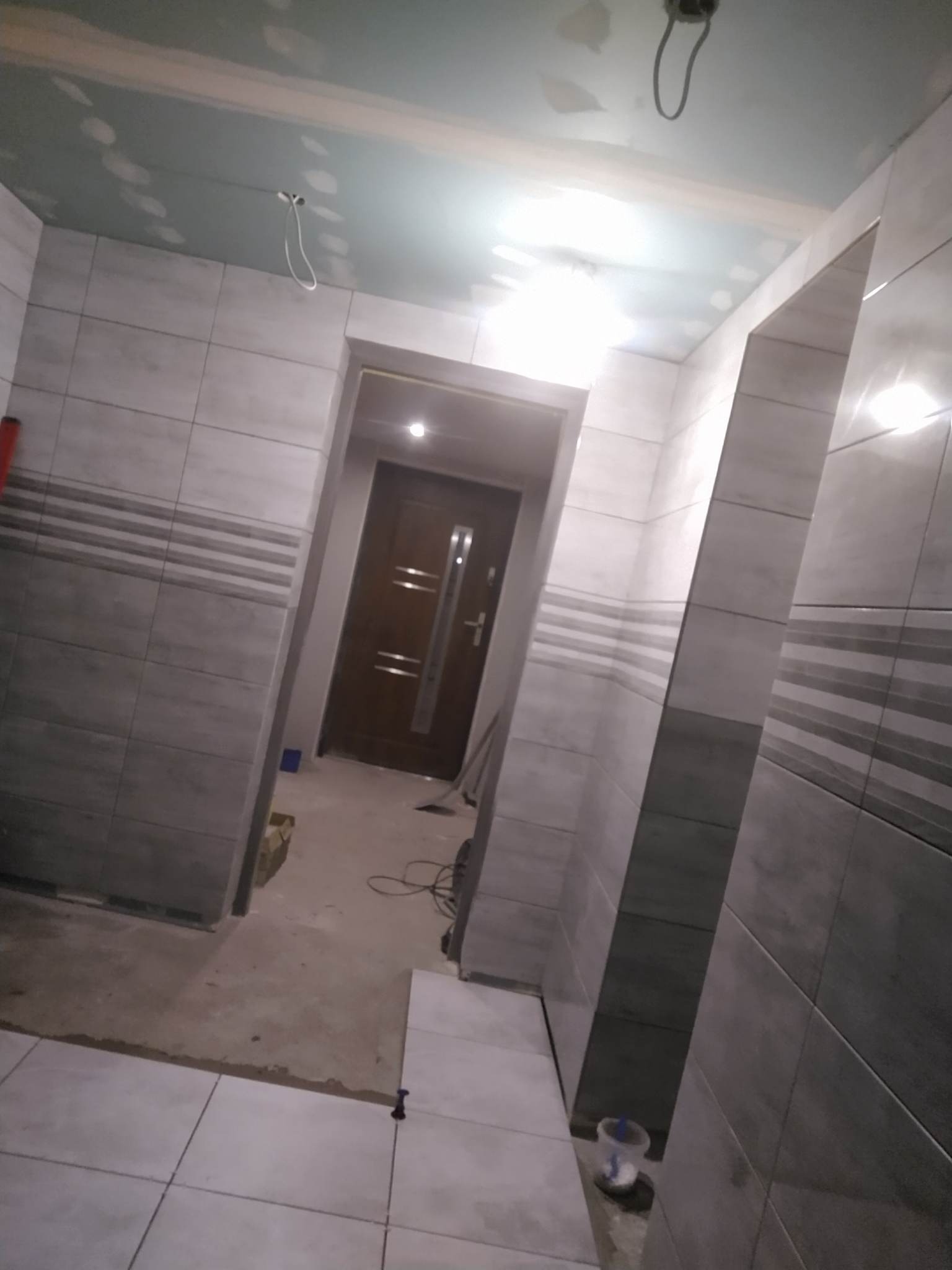 Wnętrze łazienki w trakcie remontu: ułożone szare płytki z poziomymi paskami na ścianach, sufit z płyt gipsowych, widoczne przewody elektryczne i fragment wejścia z drzwiami.