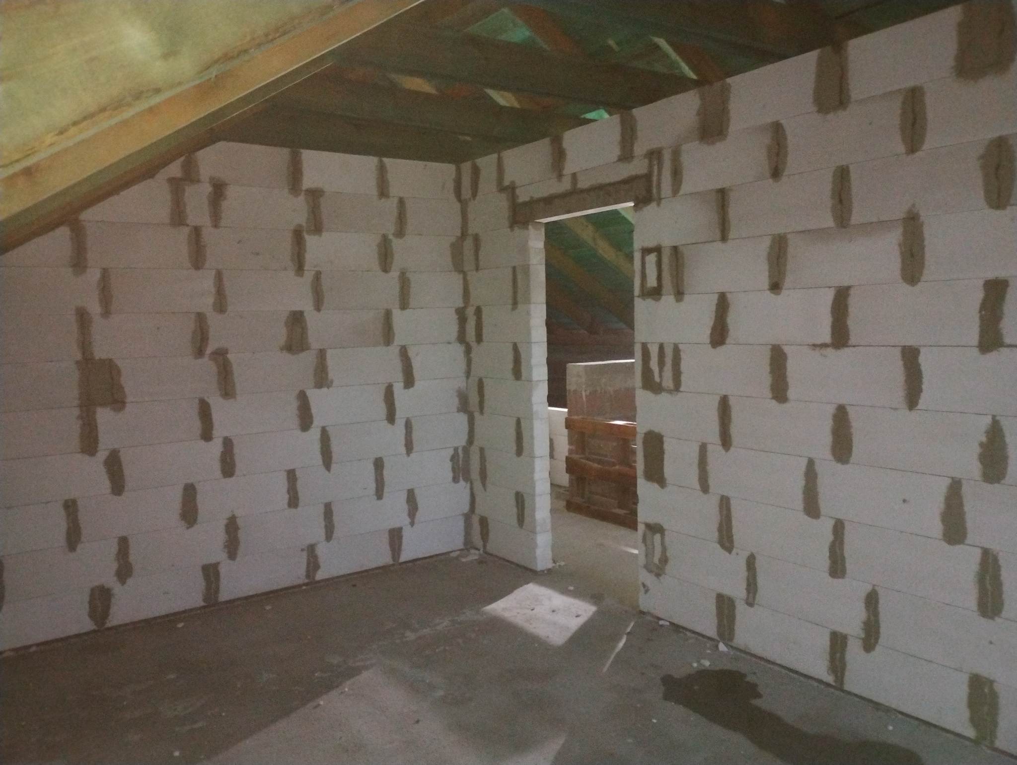 Wnętrze domu w trakcie budowy: białe ściany z betonu komórkowego z widocznymi spoinami, drewniana konstrukcja dachu, surowa podłoga, otwór drzwiowy.